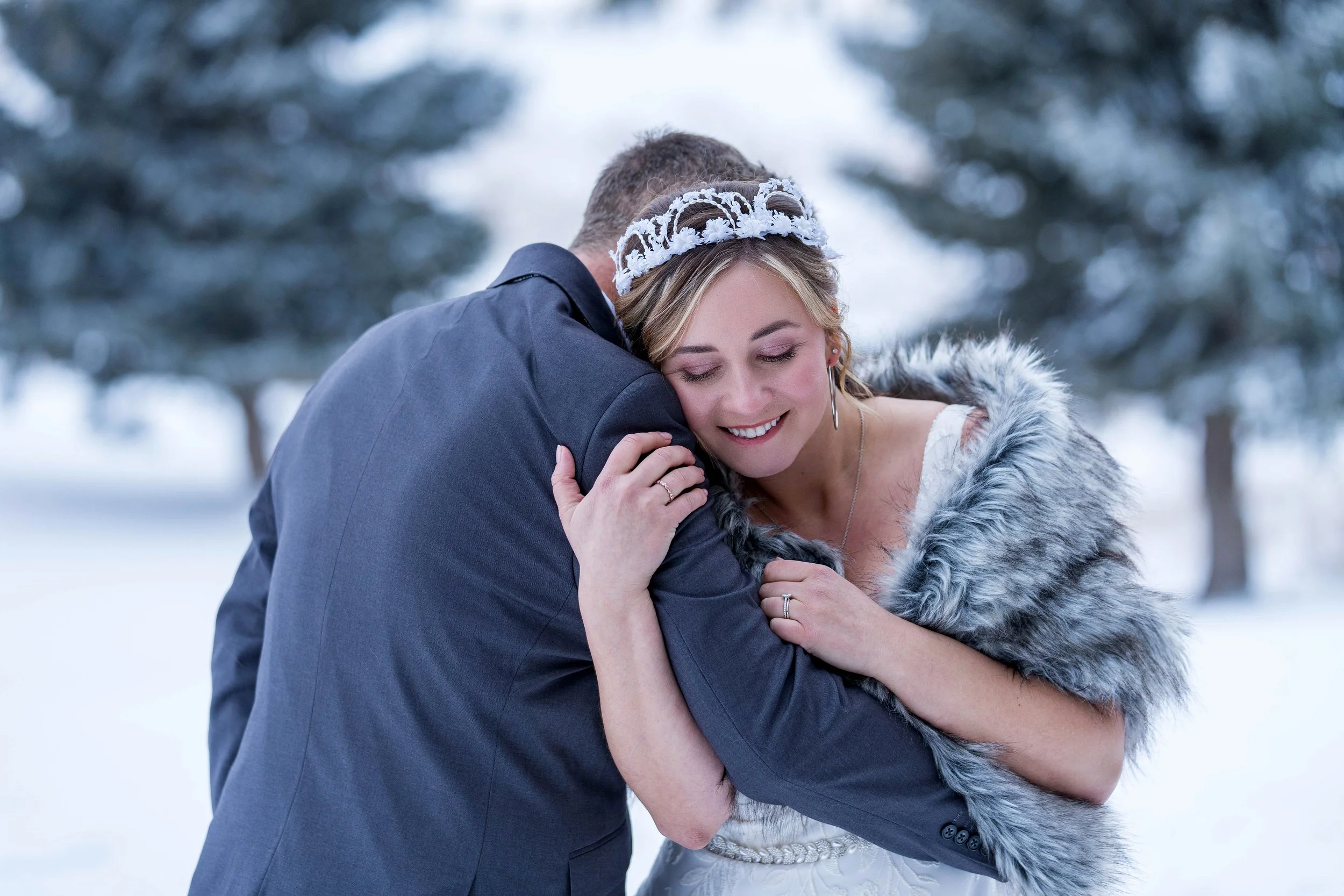 A bride and groom sharing a warm embrace outdoors in a snowy landscape, with the bride smiling and eyes closed.