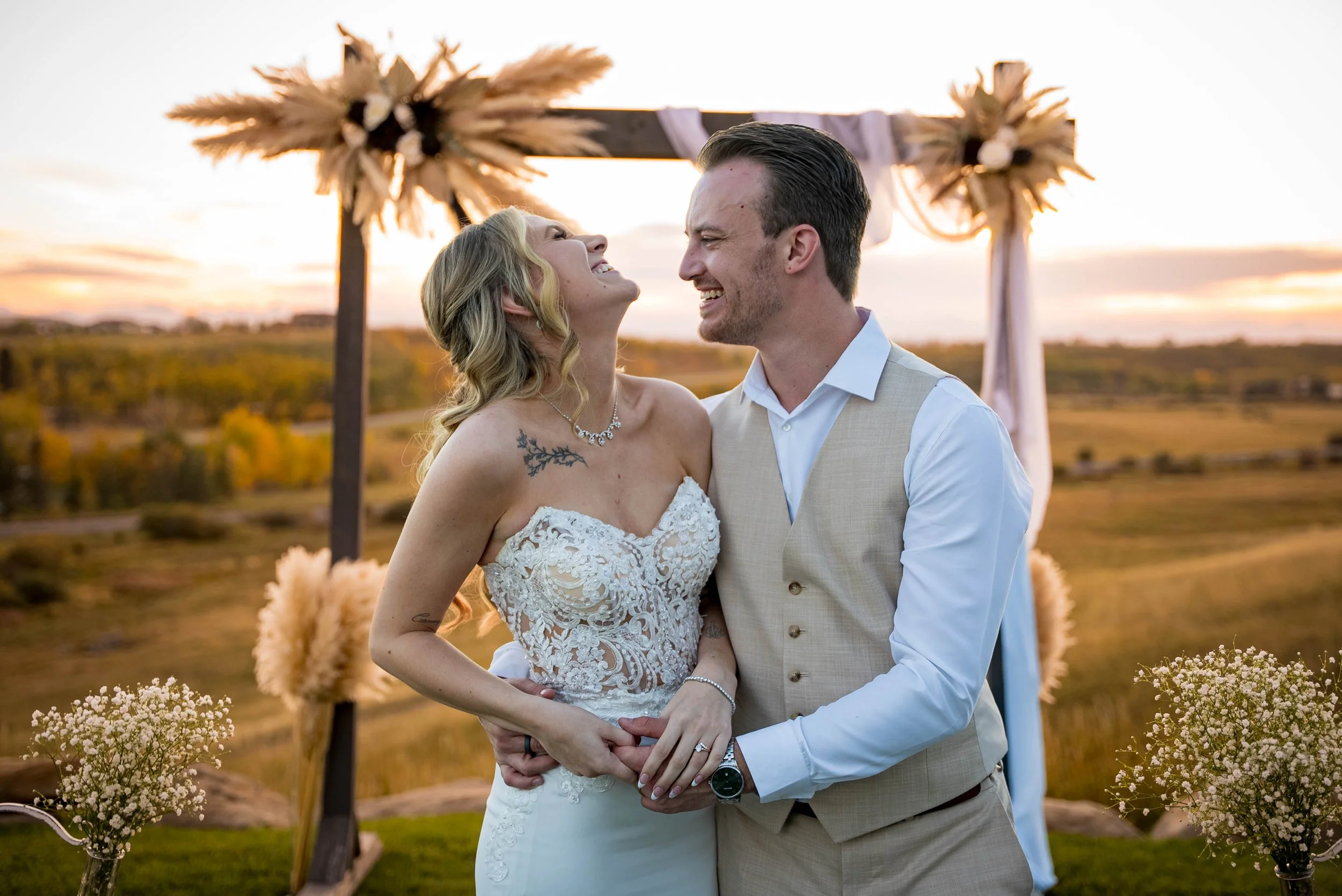 Bride and groom smiling and hugging at their outdoor wedding ceremony during sunset, with floral arrangements and a rustic arch in the background.
