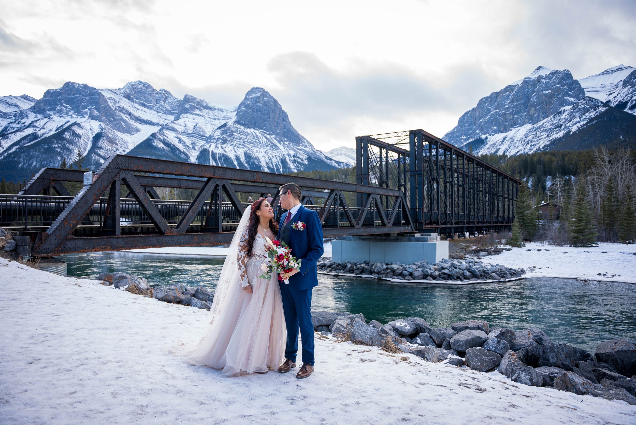 A bride and groom stand close together on snow-covered ground near a river, looking into each other's eyes, with snow-capped mountains and a train bridge in the background.