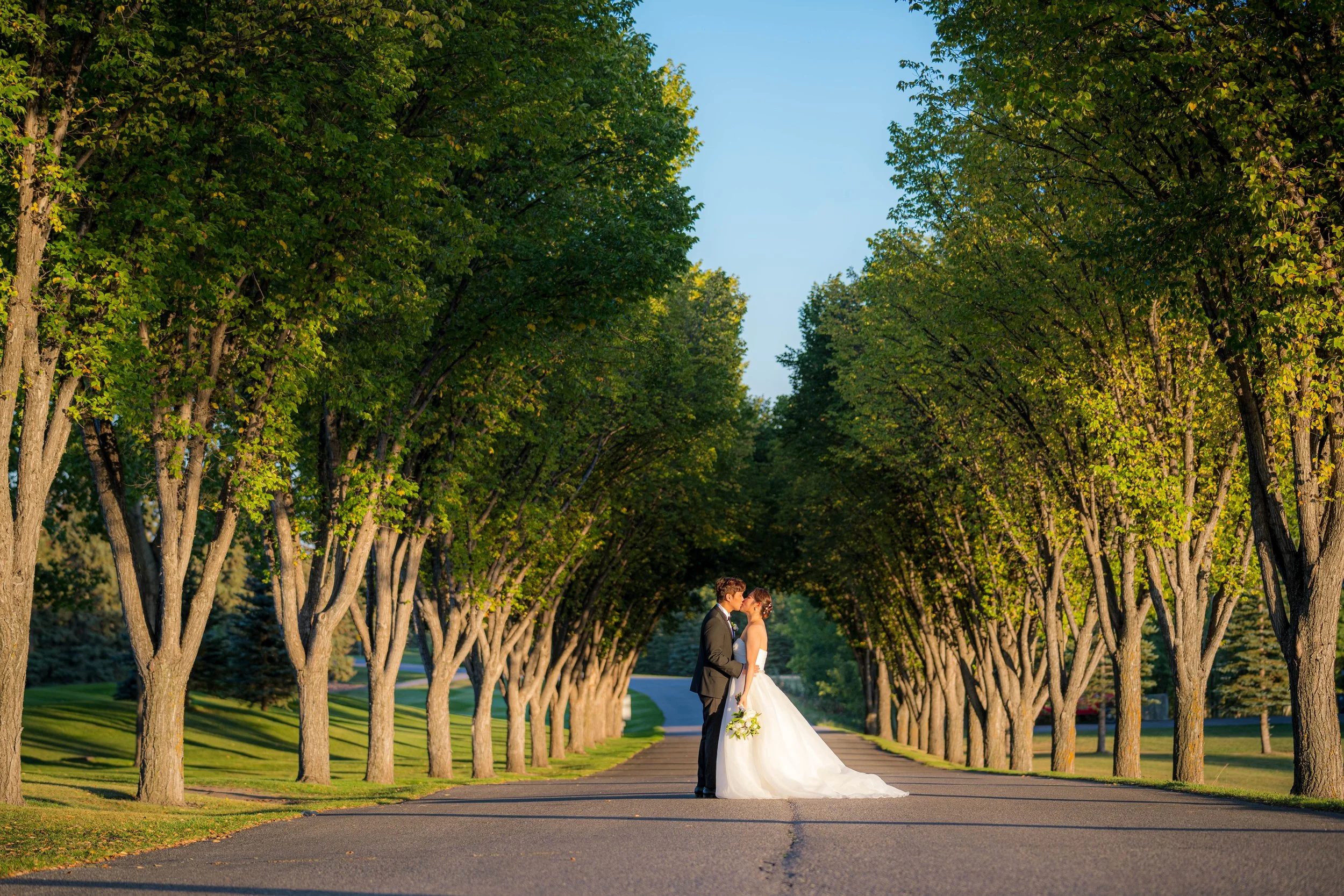 A bride and groom standing on a tree-lined road, leaning in for a kiss, with sunlight illuminating the scene.