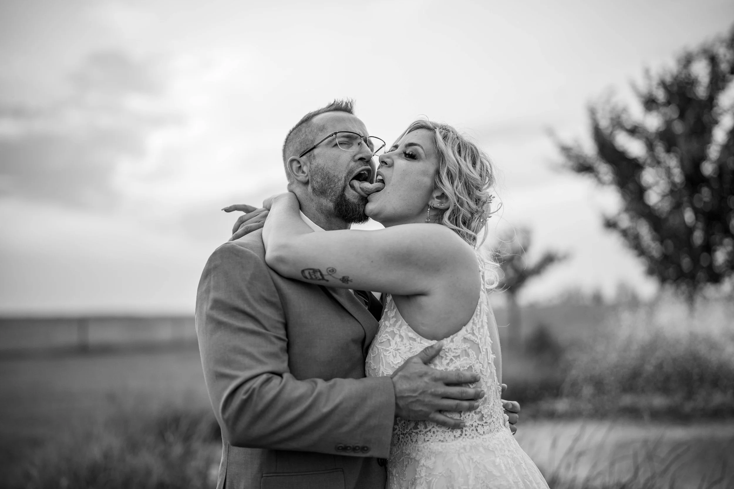 Black and white photo of a couple embracing outdoors, the woman playfully licking the man's tongue, both with closed eyes, with trees and sky in the background.