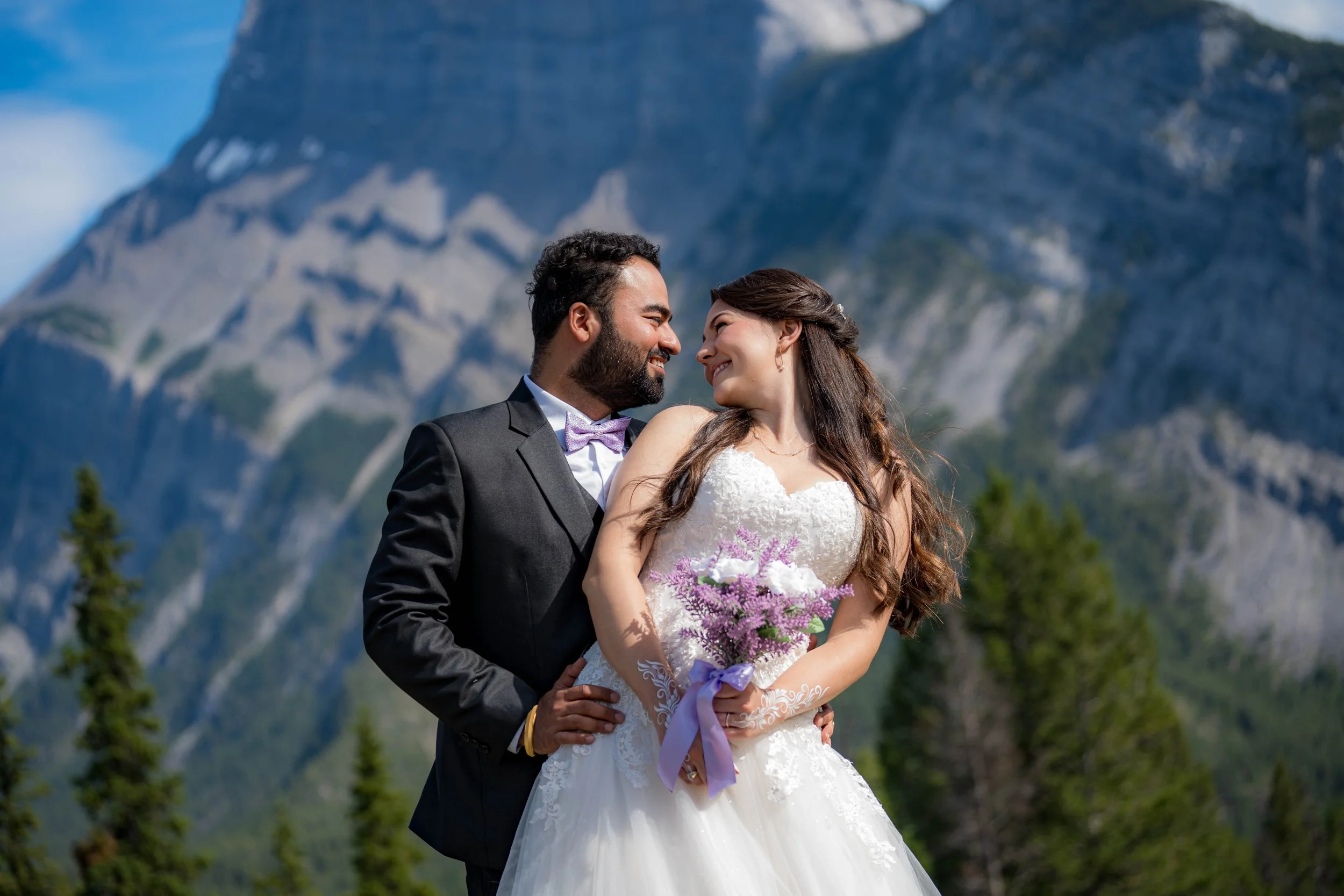 A newlywed couple gazing into each other's eyes outdoors with mountains and trees in the background. The bride is holding a bouquet of purple and white flowers, and the groom is in a black suit with a purple bow tie.