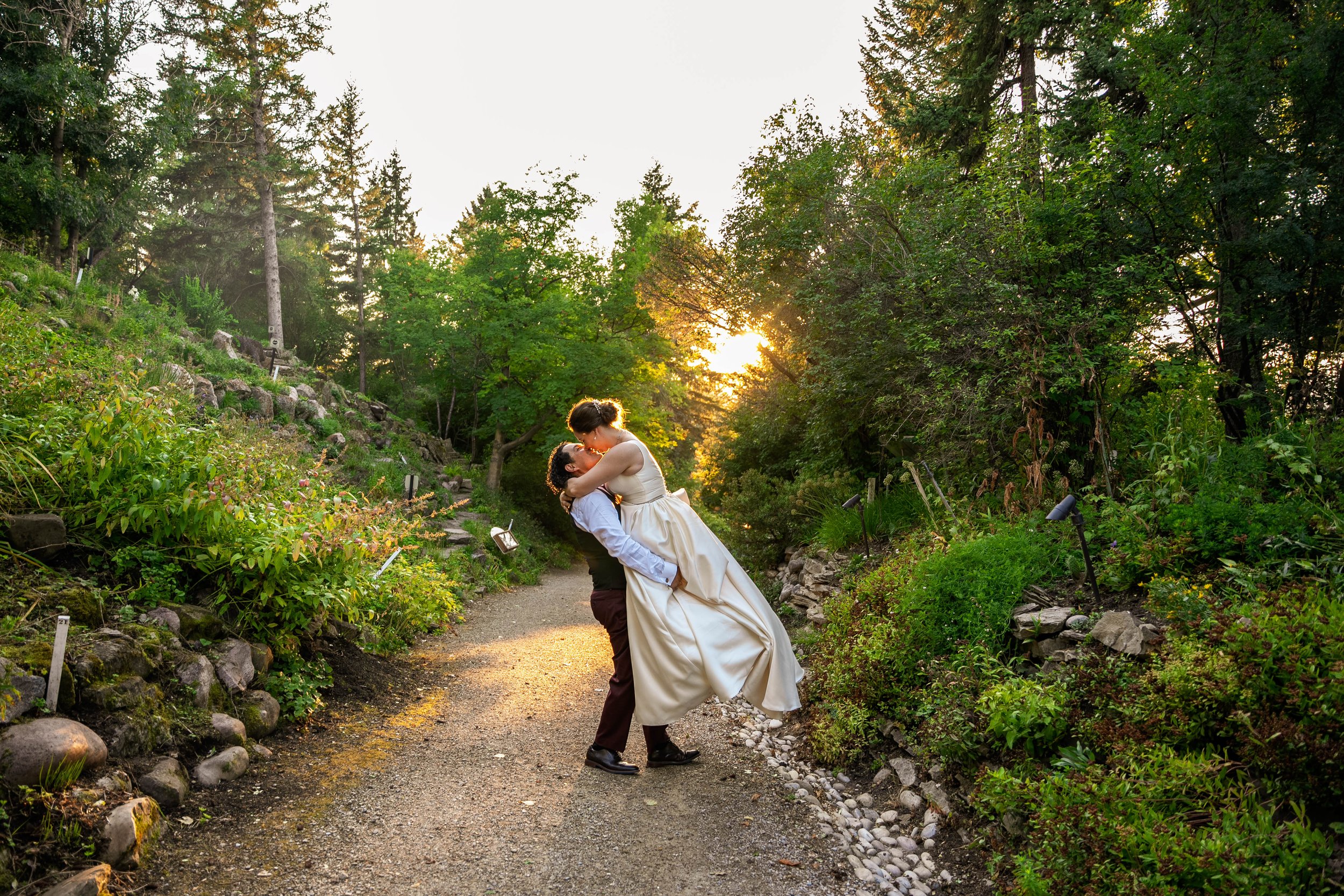A couple dances in a garden path during sunset, surrounded by trees and greenery.