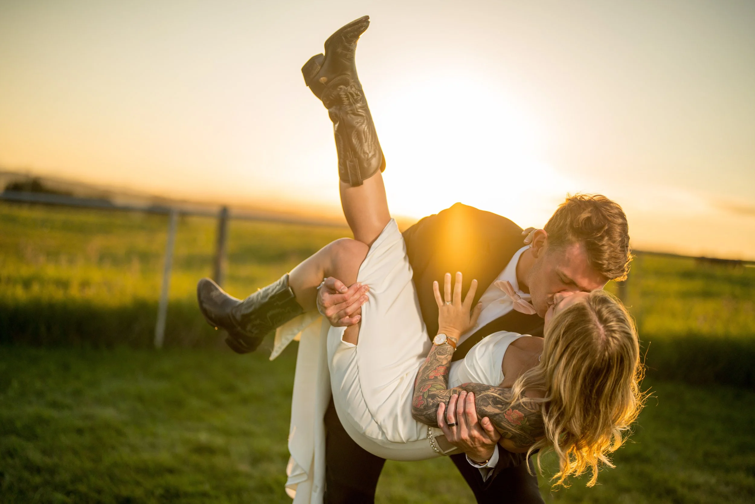 A couple dressed in formal wedding attire dancing and kissing outdoors during sunset, with the groom holding the bride in his arms.