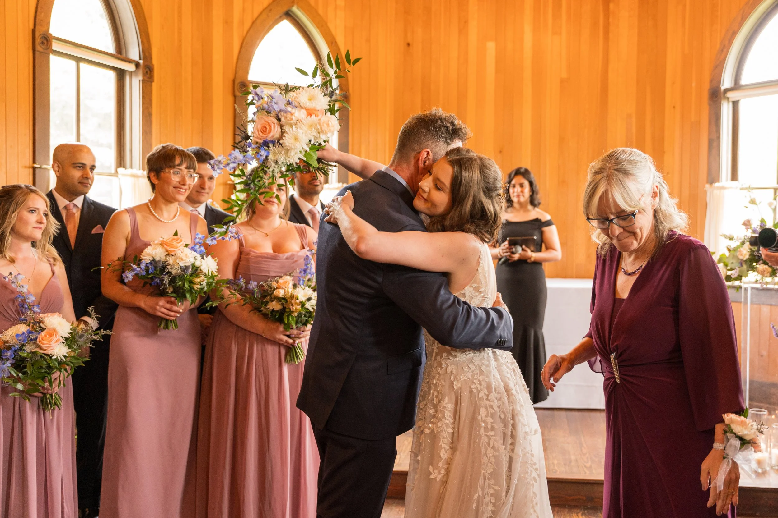 A bride and groom hugging during a wedding ceremony inside a wooden chapel, with bridesmaids and groomsmen holding bouquets of flowers standing nearby, and an officiant standing to the right.