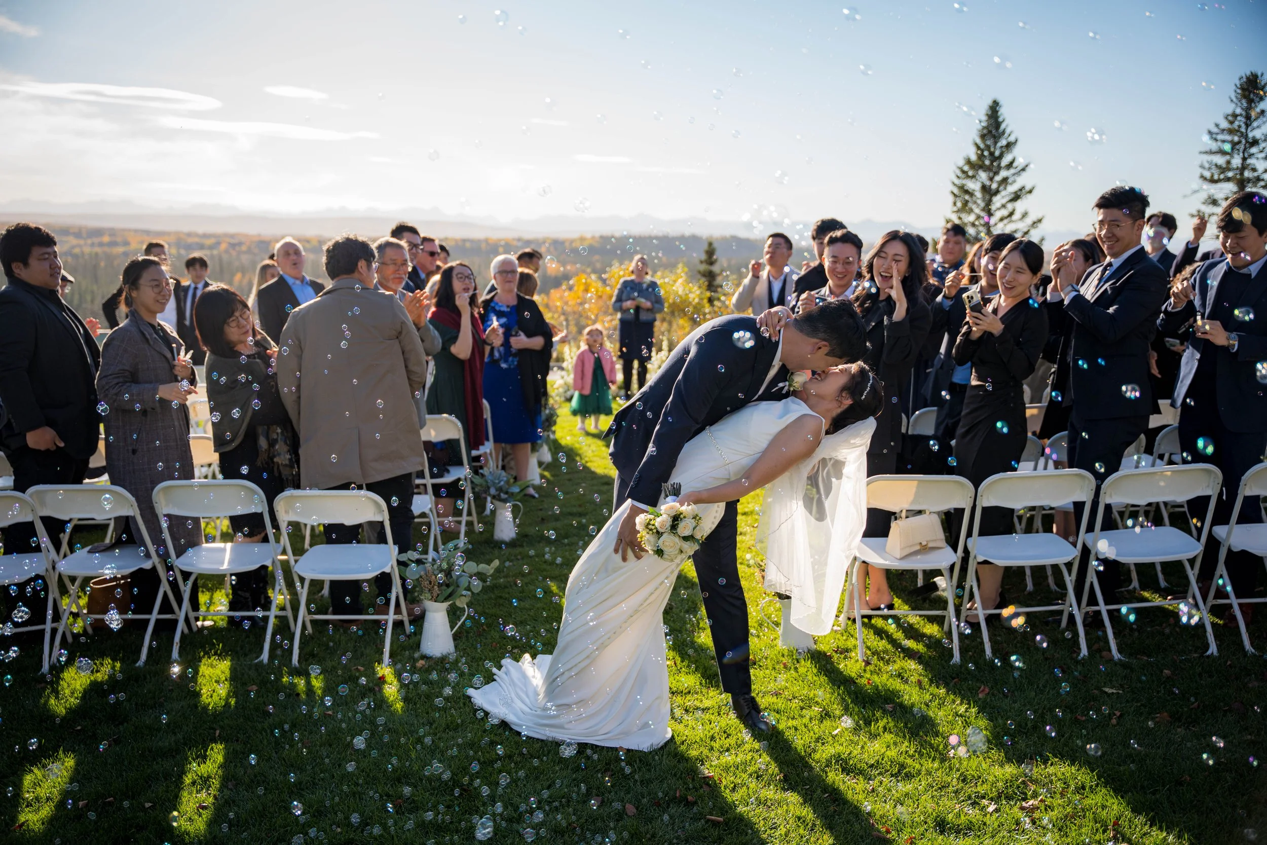 A newlywed couple sharing a kiss outdoors at their wedding, surrounded by guests and bubbles, with sunlight and trees in the background.