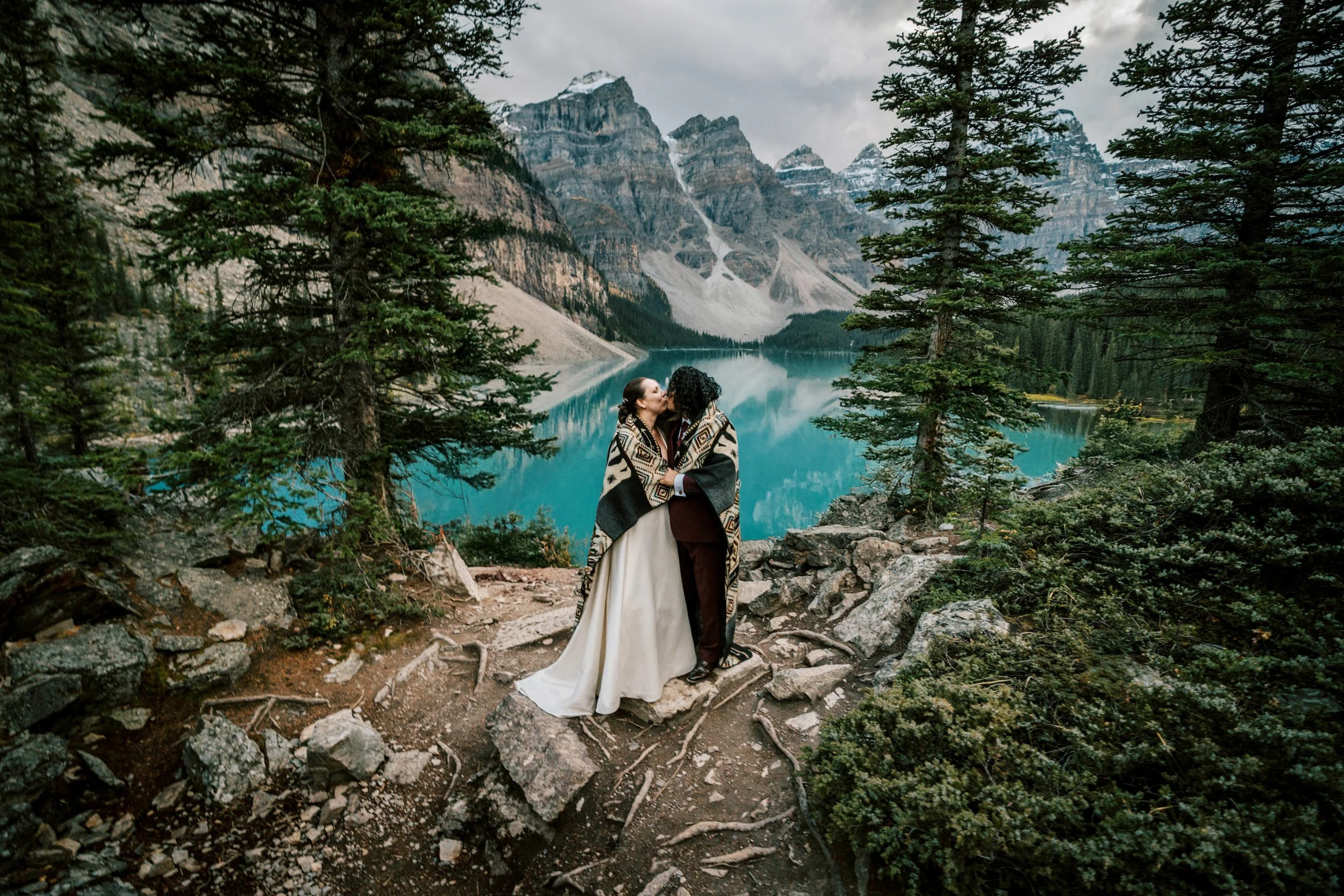 Couple sharing a kiss on a rocky path near a mountain lake, surrounded by evergreen trees with snow-capped mountains in the background.