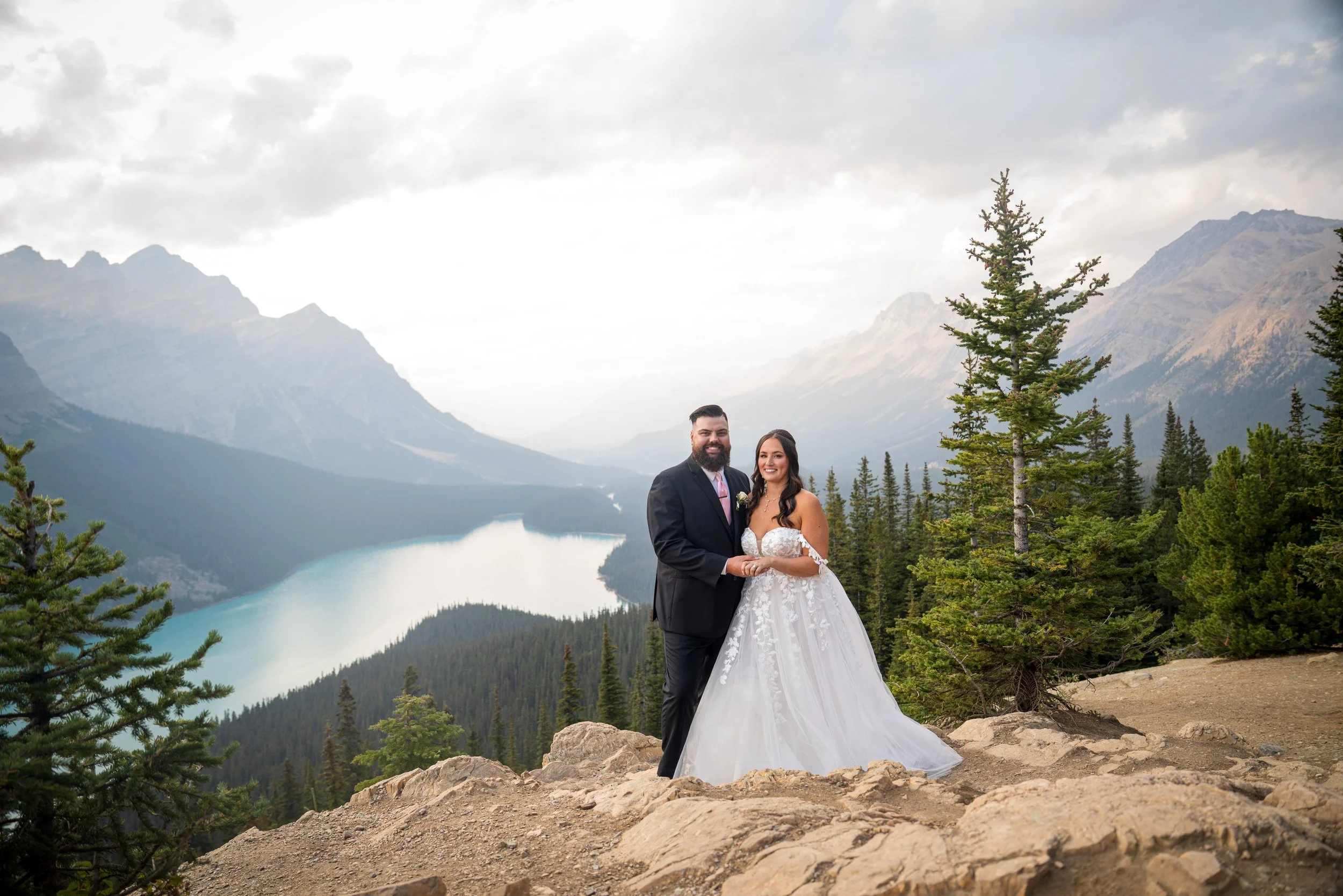A bride and groom standing hand in hand on a rocky ledge with a mountain lake, evergreen trees, and mountains in the background.