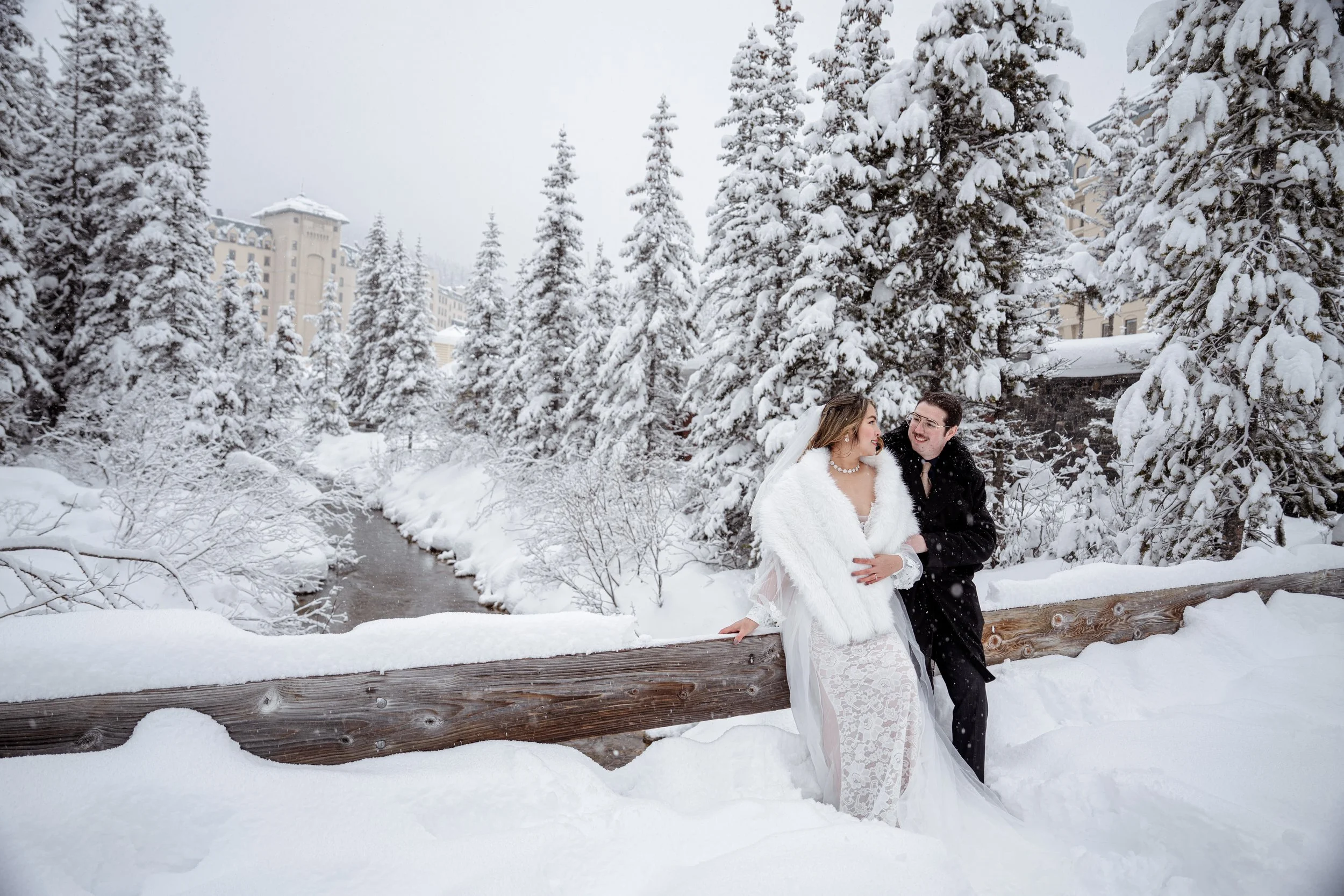 A bride and groom stand outdoors in a snowy landscape, with snow-covered trees and a building in the background. The bride wears a white wedding dress with a fur shawl, and the groom wears a black suit. They are smiling and looking at each other, lea