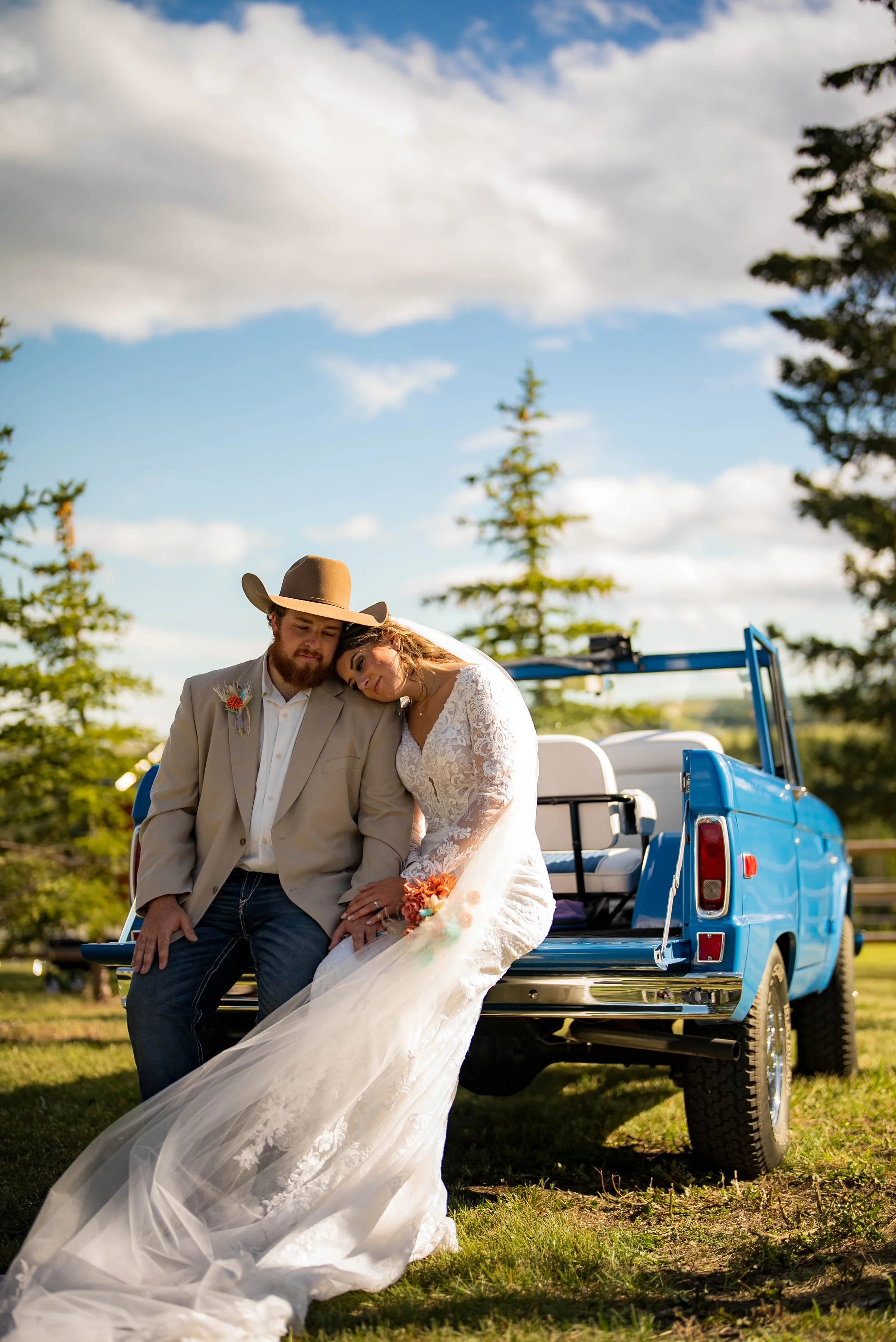 Bride and groom sitting on the tailgate of a blue pickup truck with a scenic outdoor background, embracing and resting their heads together on a sunny day.