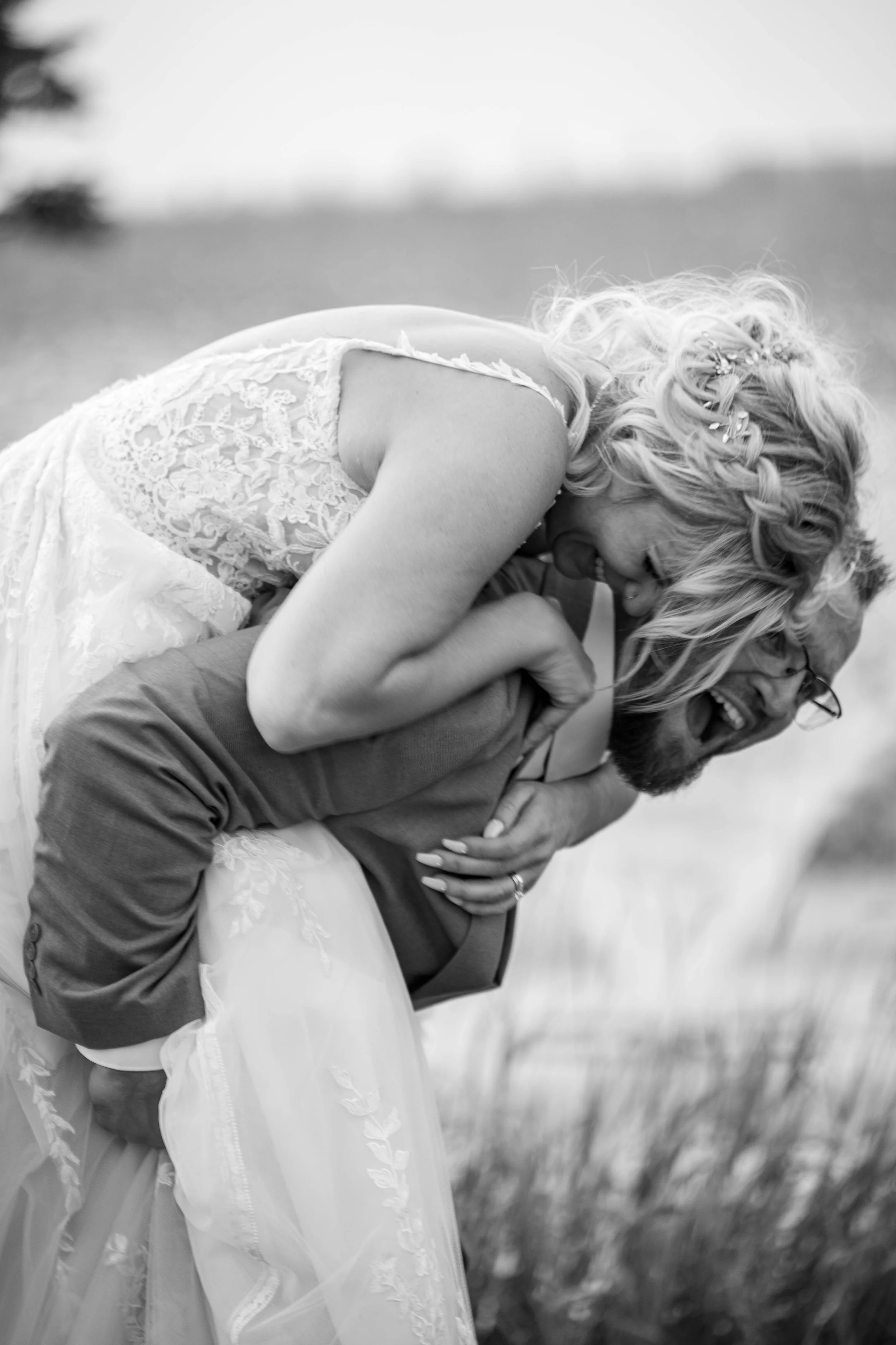 Black and white photo of a woman in a wedding dress and a man in a suit, playfully interacting outdoors with tall grass and trees in the background.