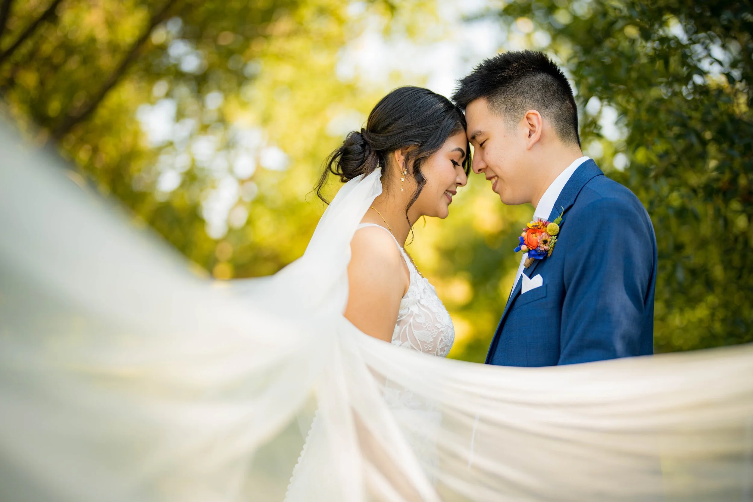 A bride and groom with foreheads touching, smiling, outdoors surrounded by trees, during a wedding photoshoot.