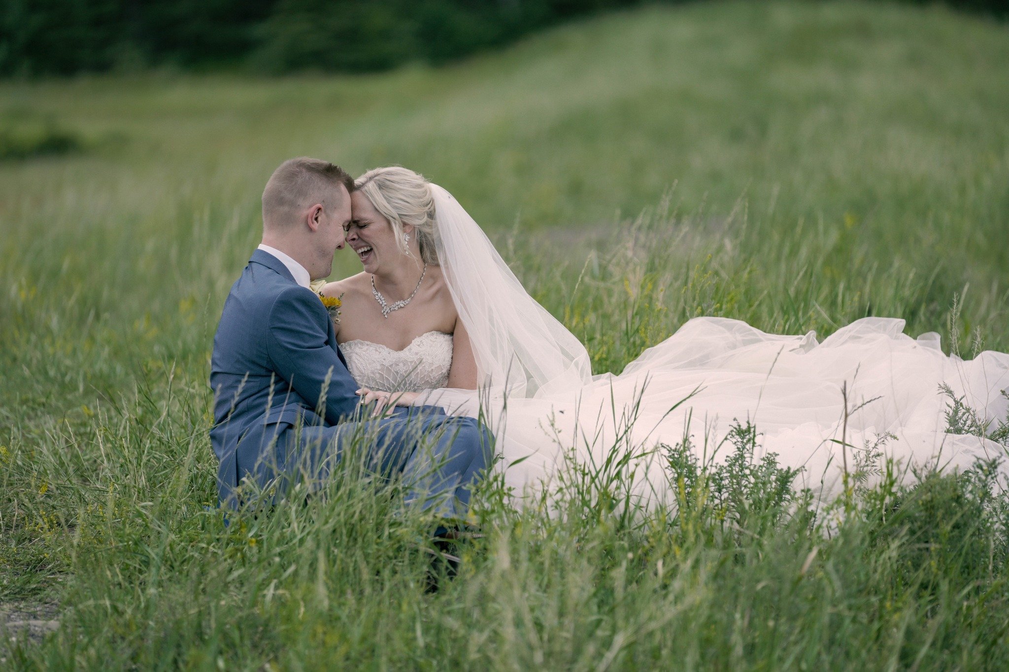 A bride and groom sitting in a grassy field, laughing and enjoying a joyful moment on their wedding day.