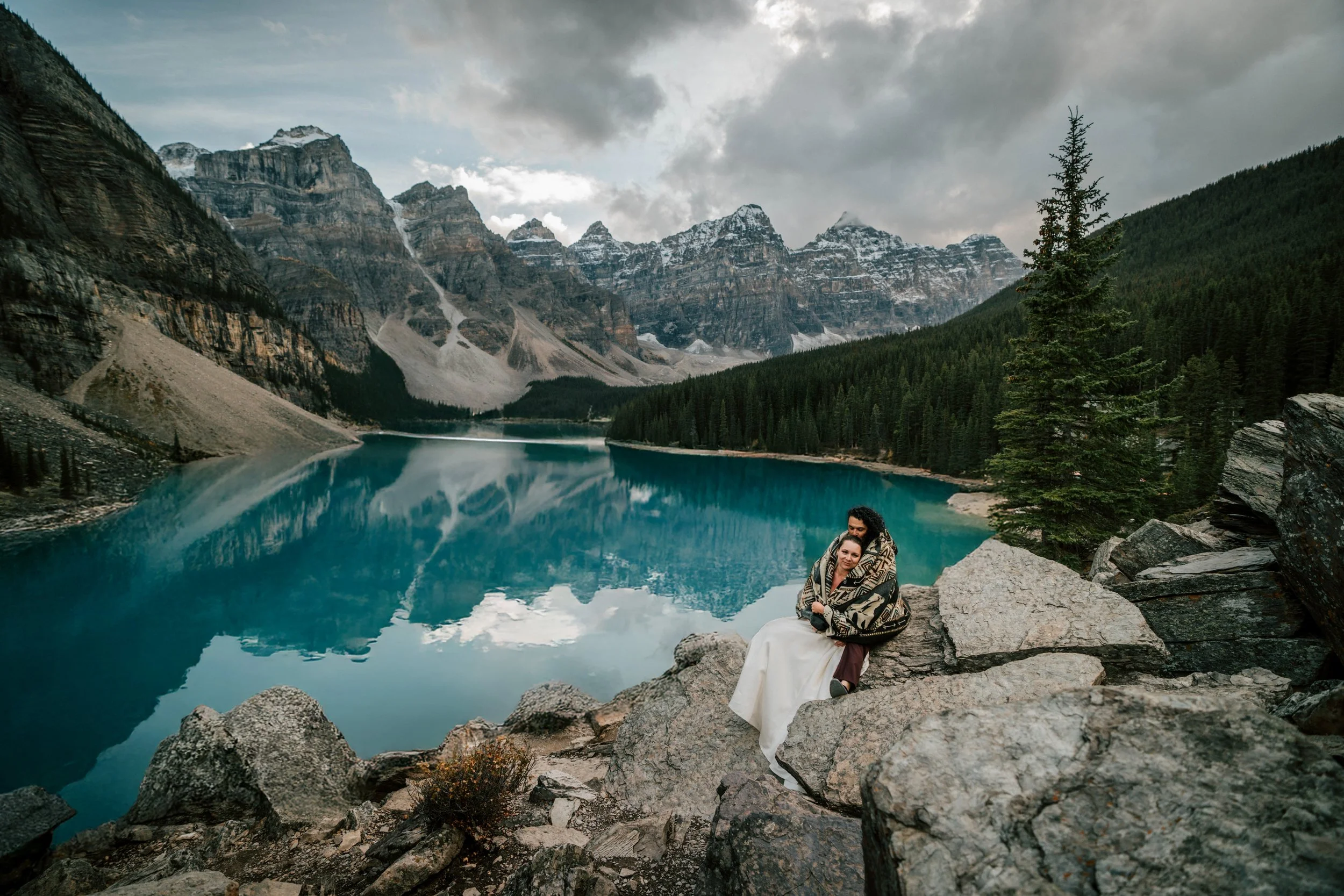 Oscar and Michaela wrapped in a blanket sitting together on the rocks overlooking Moraine Lake Banff National Park with full turquoise lake and mountain reflection