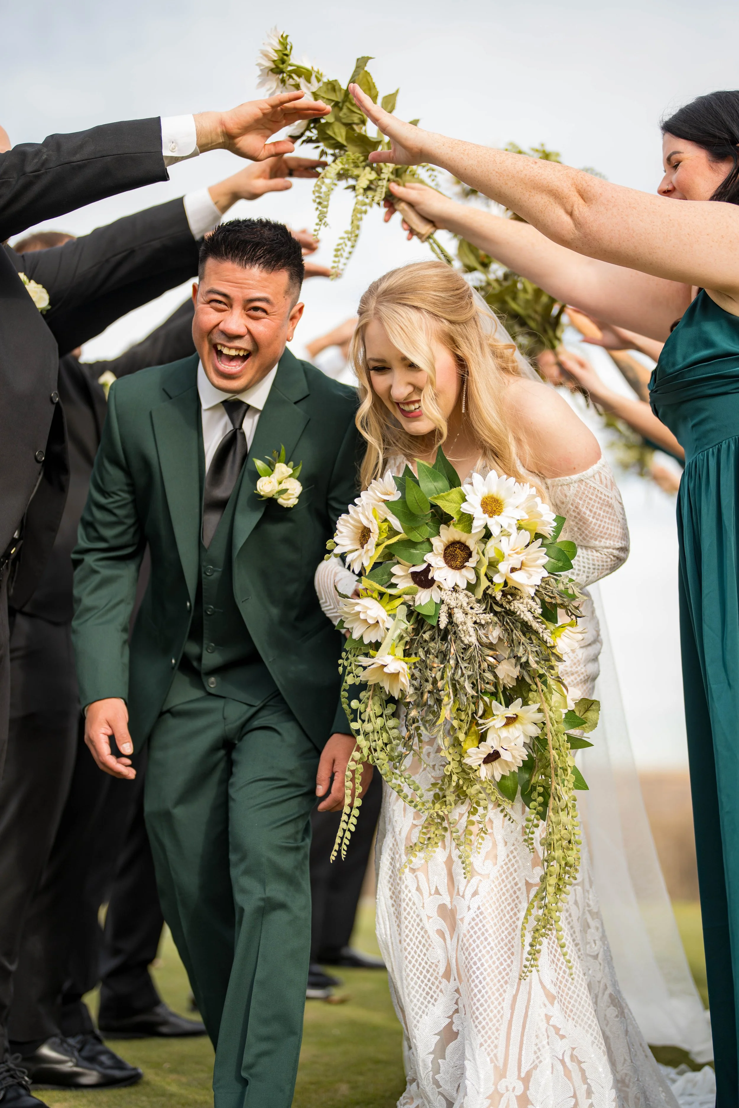 Happy bride and groom walking under a floral arch at an outdoor wedding, surrounded by guests in formal attire.