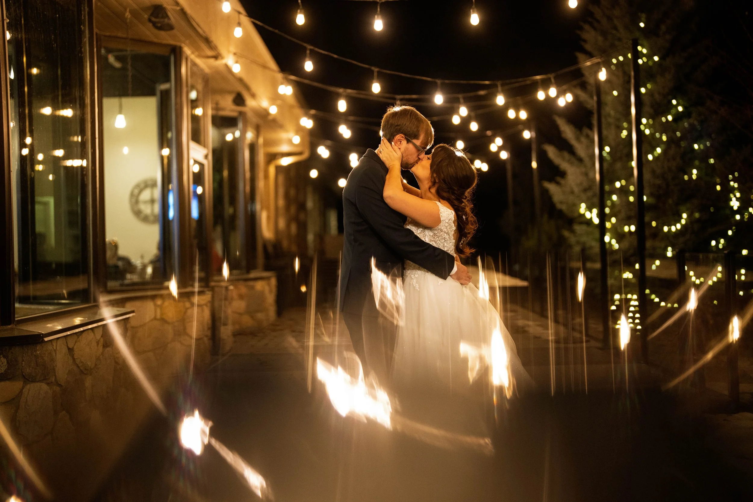 A couple in wedding attire sharing a kiss and embrace under string lights on a night outdoor patio.