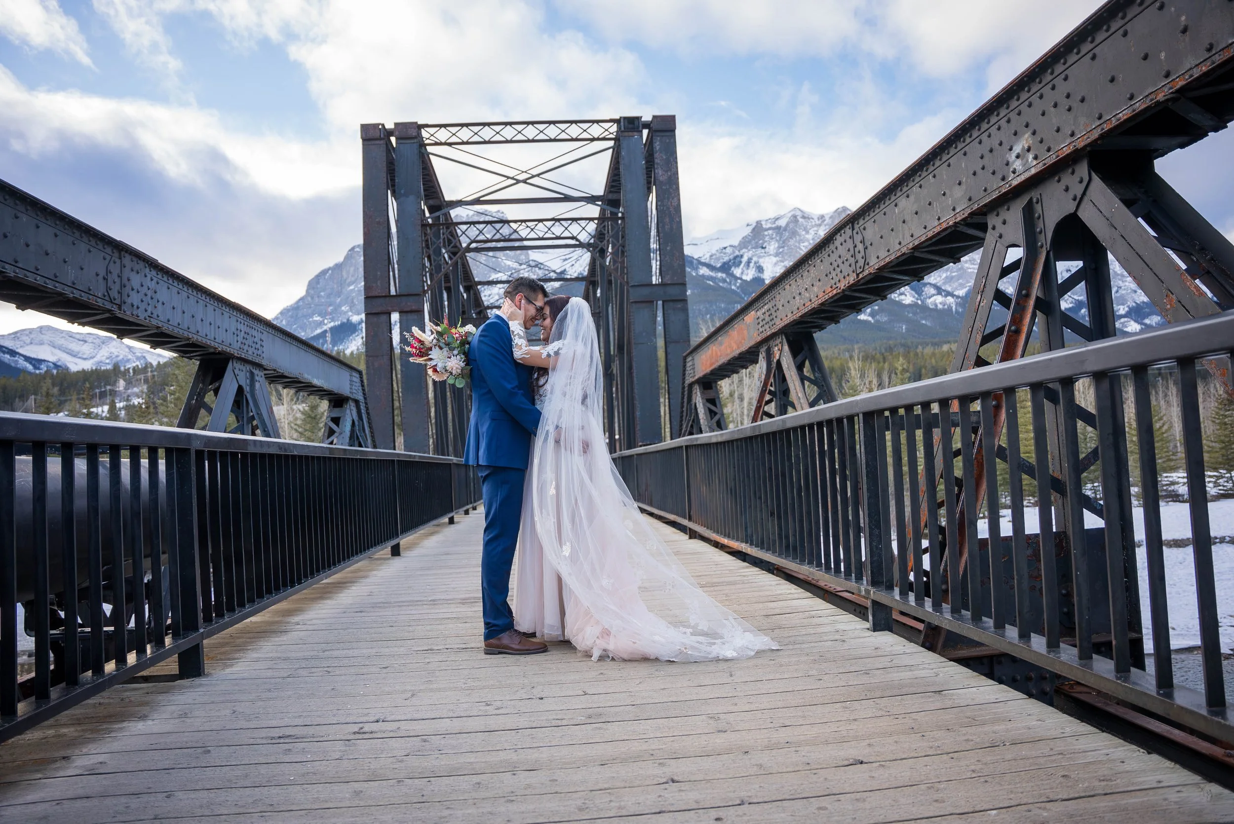 A bride and groom in wedding attire sharing a kiss on a wooden bridge with mountain scenery in the background.