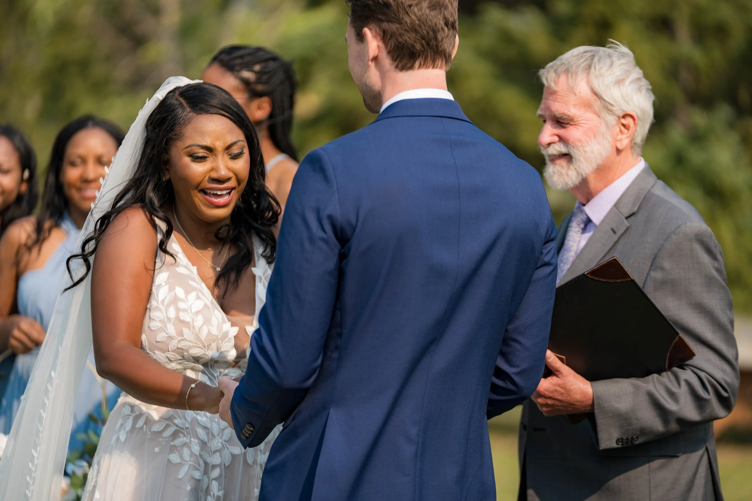 A bride crying during her wedding ceremony as a groom holds her hands, with an officiant smiling nearby and bridesmaids in the background.
