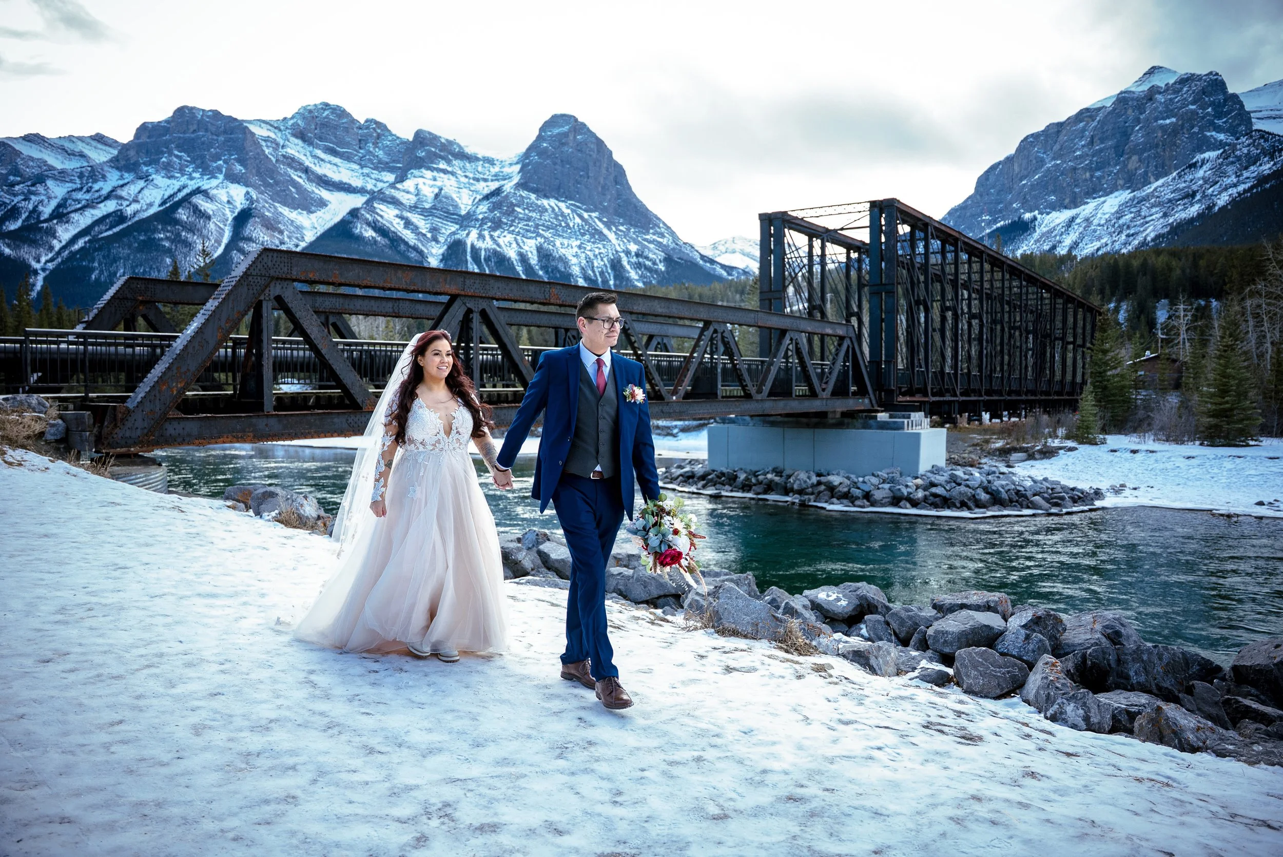 A bride and groom holding hands and walking along a snowy riverbank with mountains in the background. The bride is wearing a white wedding dress and a veil, and the groom is dressed in a blue suit and carrying a bouquet.