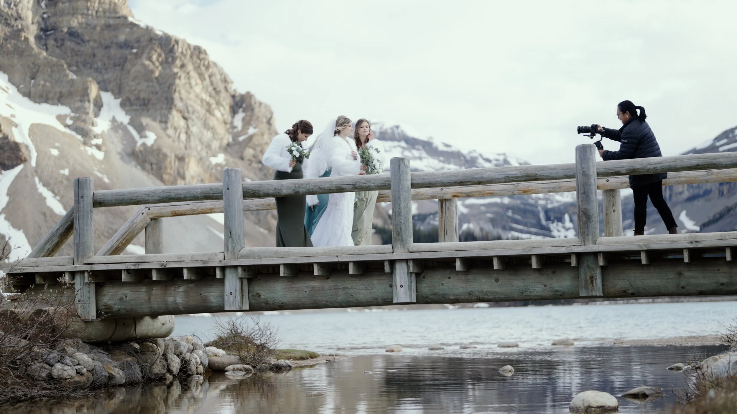Wedding ceremony on a wooden bridge over a mountain lake with snow-capped mountains in the background. Three women, one in a wedding dress, stand together, while a photographer takes pictures.