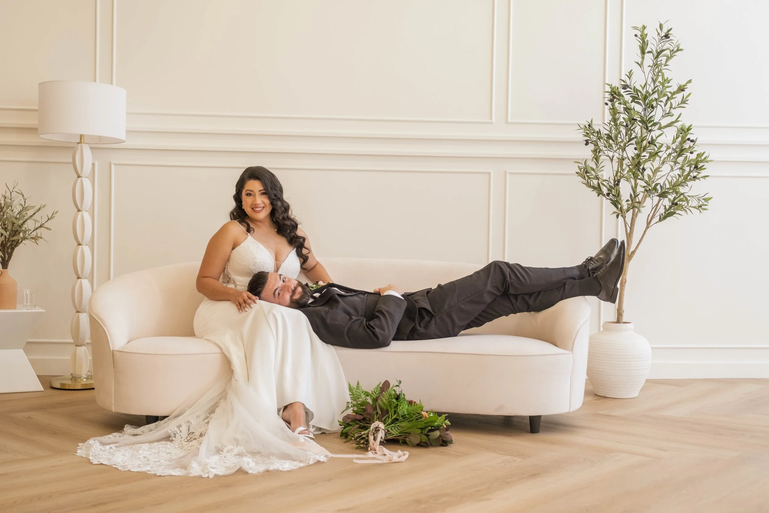Couple on a white couch in a minimalist room, woman in a white wedding dress sitting, man in a black suit lying with legs extended, bouquet on the floor, potted plants and lamps nearby.