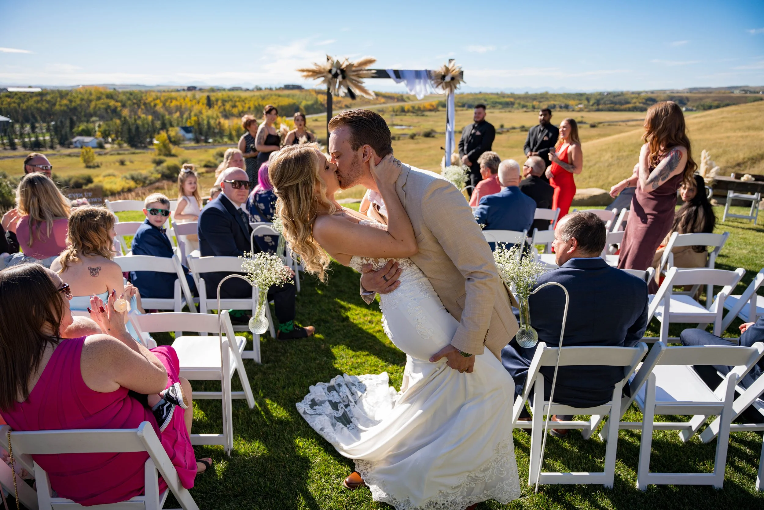 A newlywed couple shares a kiss at their outdoor wedding ceremony, with guests seated around them and a scenic landscape in the background.