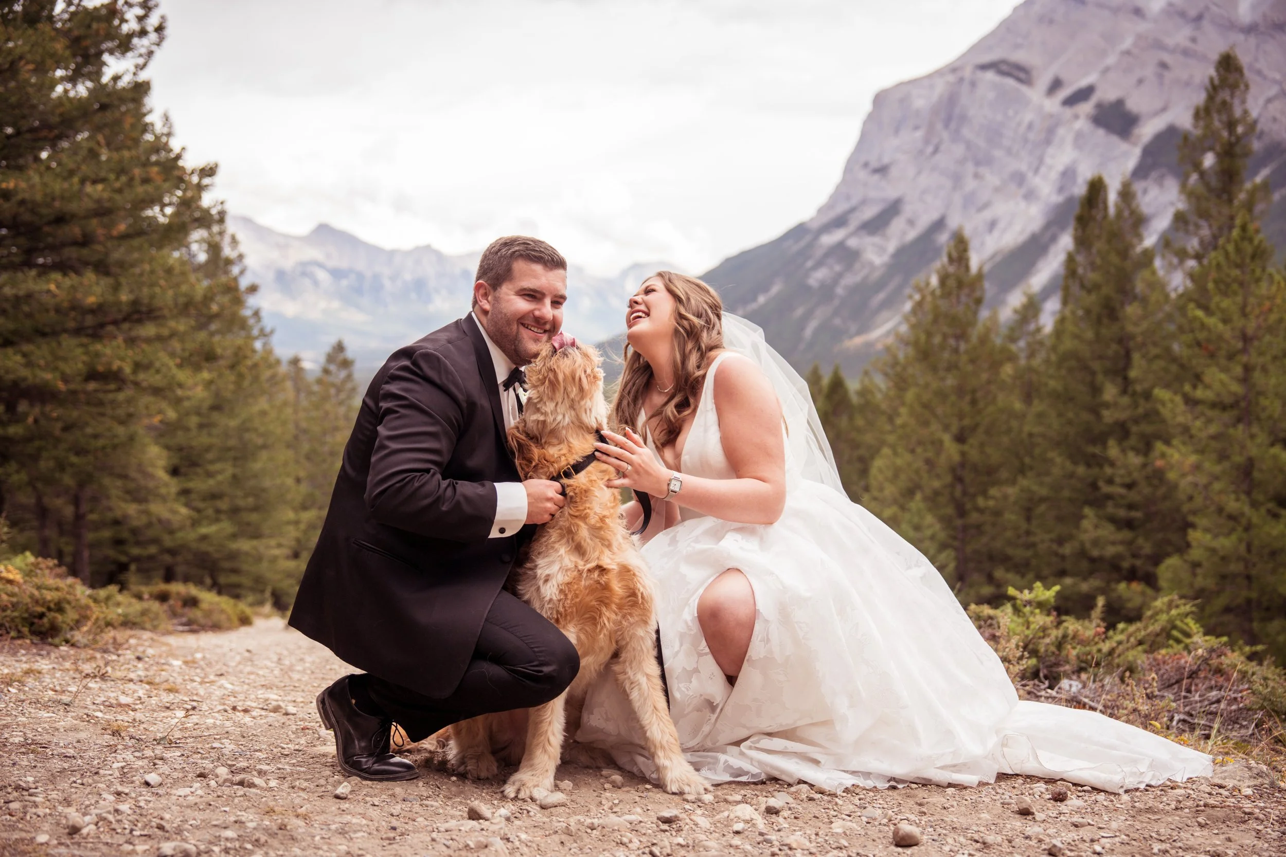 A bride and groom with their dog outdoors in a mountainous forest setting, smiling and looking at each other.