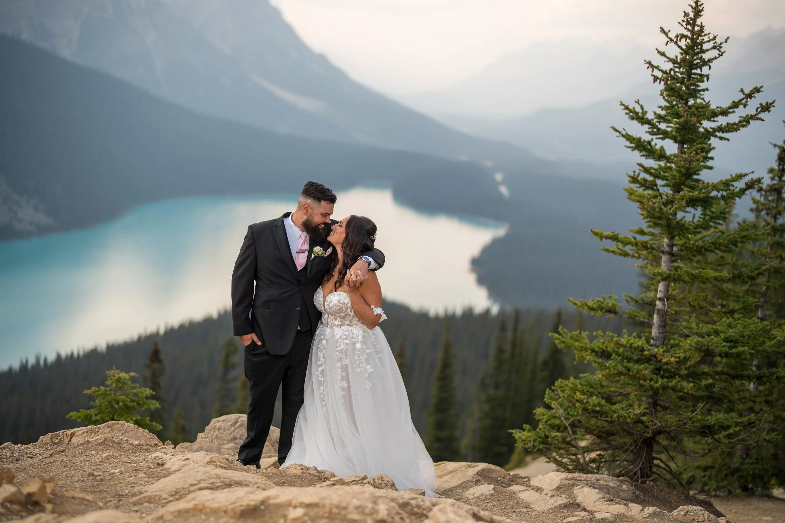 A bride and groom standing together on rocky terrain with mountains, a lake, and pine trees in the background, celebrating their wedding.