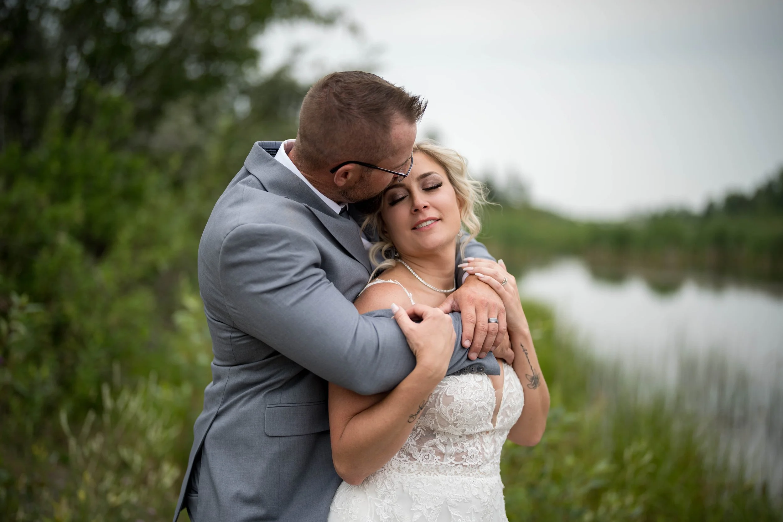 A man in a gray suit embraces a woman in a white lace dress outdoors near a body of water, with greenery in the background.
