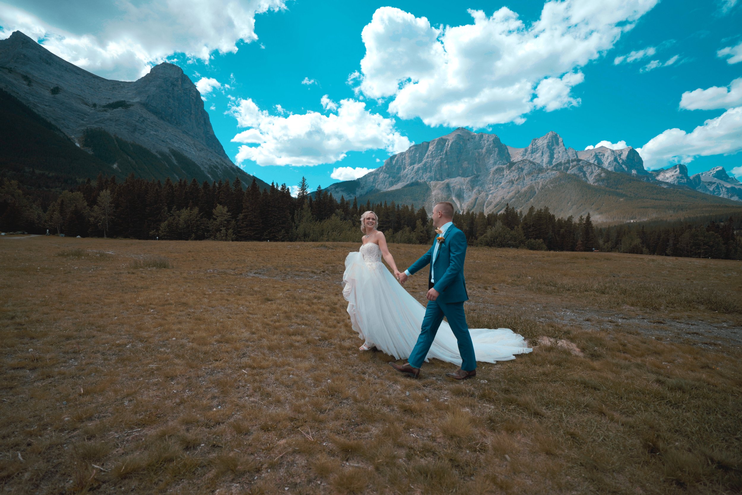 A bride and groom holding hands and walking in a field with mountains and blue sky in the background.