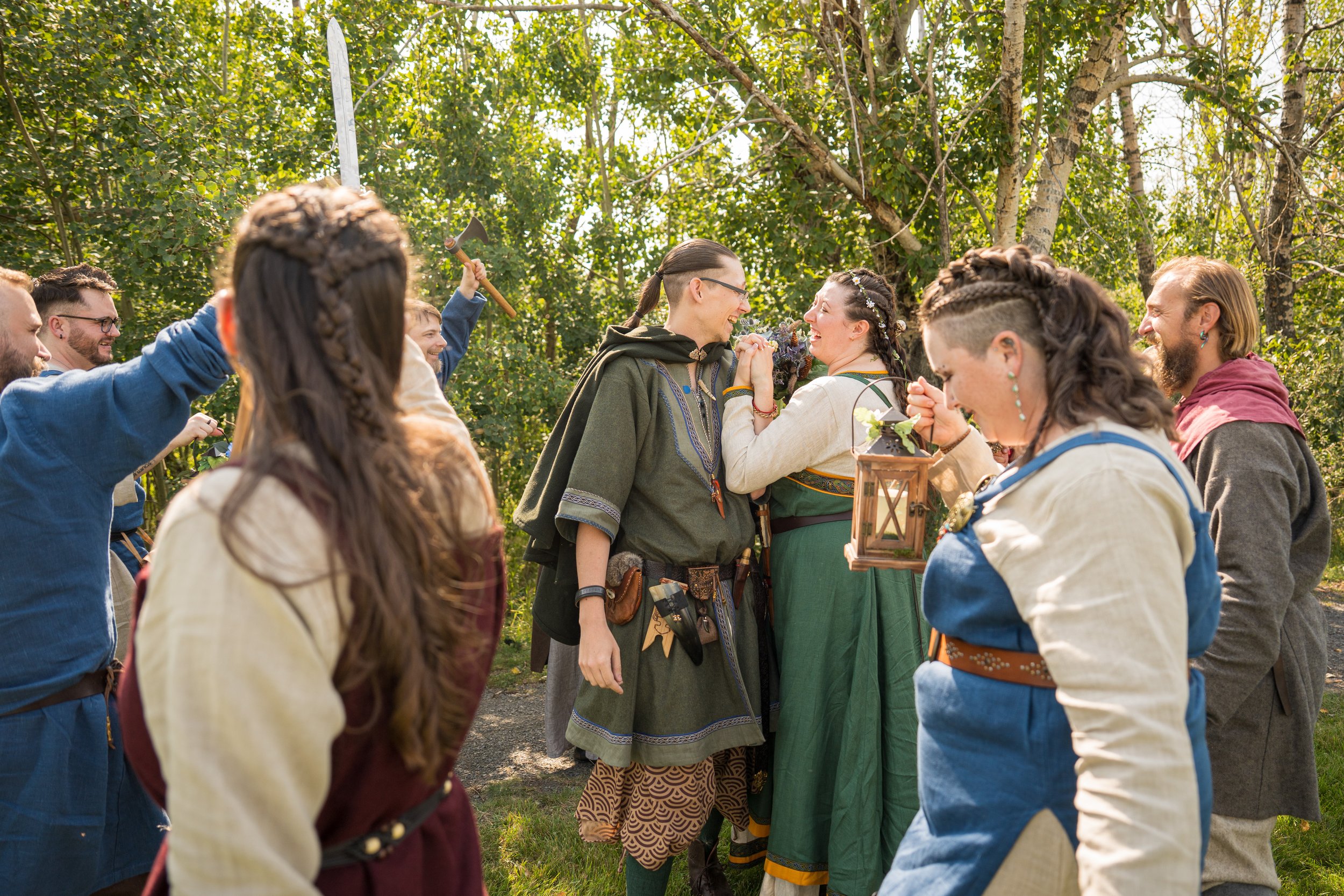 Group of people dressed in medieval costumes, celebrating outdoors among trees, with two women smiling and holding hands in the center, one wearing a green dress and the other a cream-colored dress, surrounded by friends cheering and holding props li