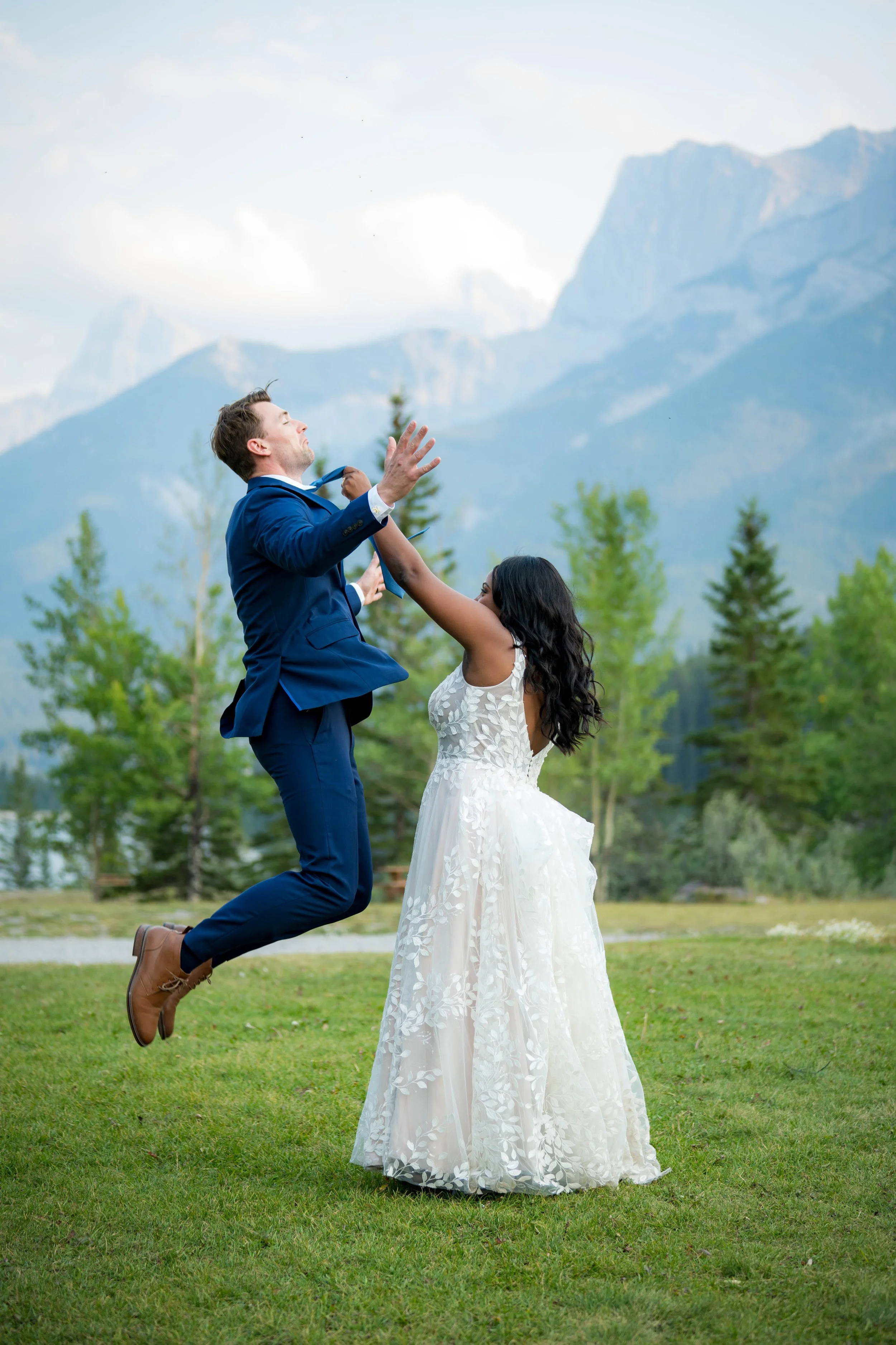 A bride and groom are outside on a grassy field with mountains and green trees in the background. The groom, in a blue suit, is jumping with his eyes closed and one arm raised. The bride, in a white lace wedding dress, is reaching out towards him.