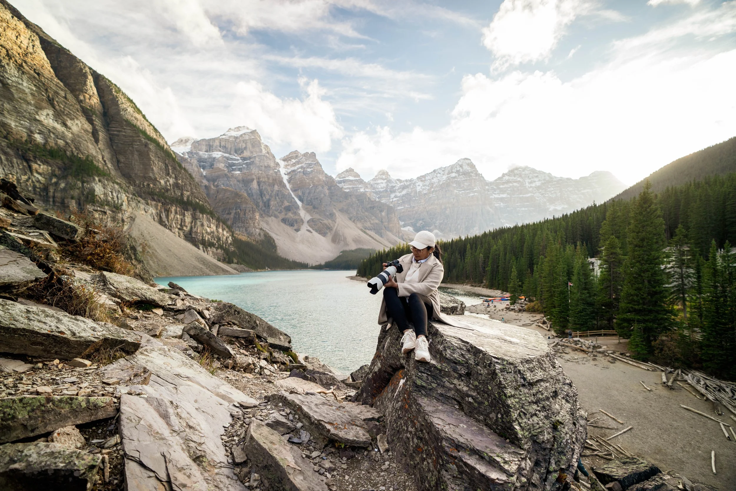 A woman in a hat and coat sitting on a large rock by a lake with mountains in the background, holding a camera or telescope, in a scenic outdoor area.