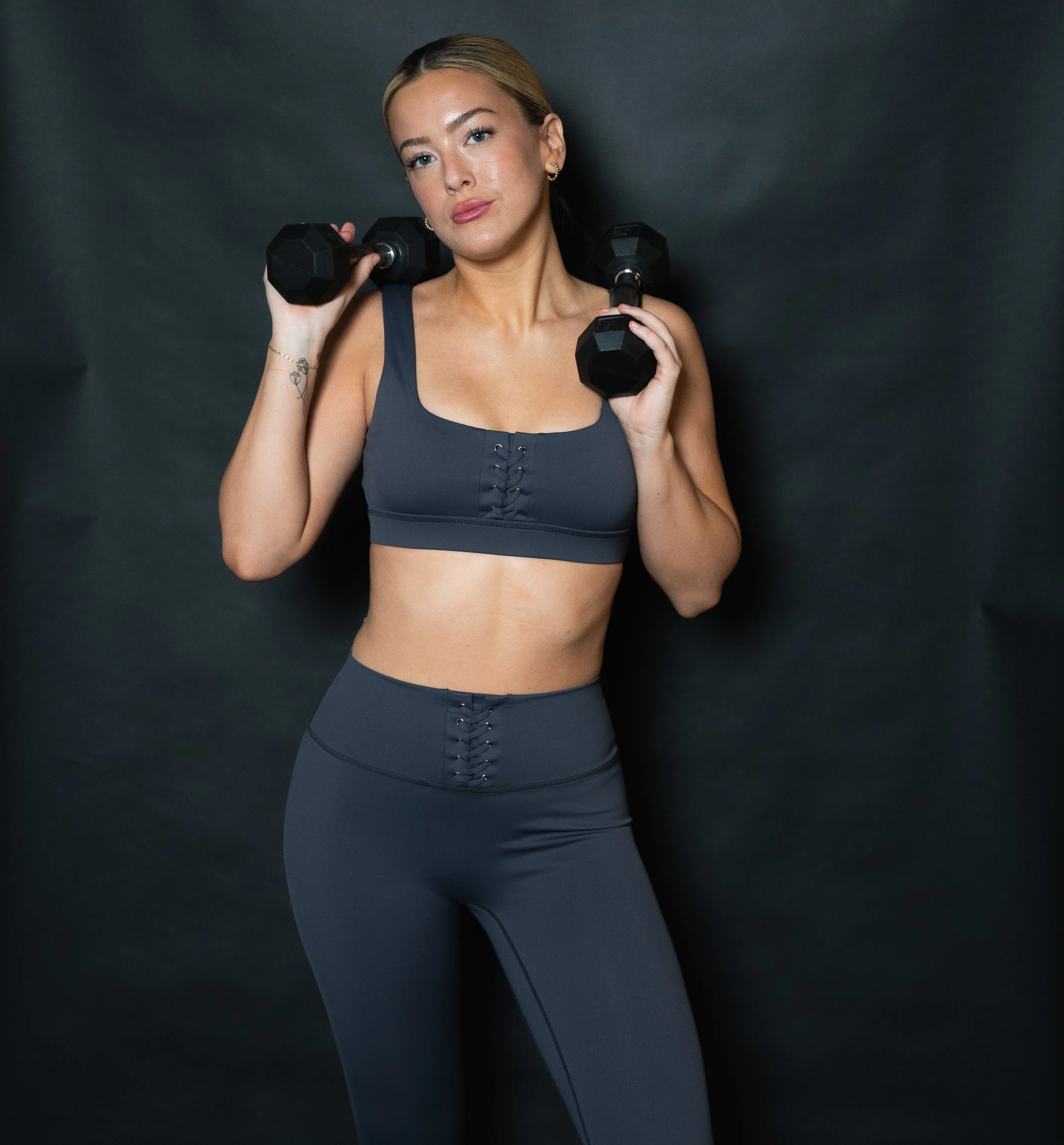 A woman with blonde hair in a ponytail wearing the melanie set,  sports bra and matching high-waisted leggings holding two dumbbells against a dark background.