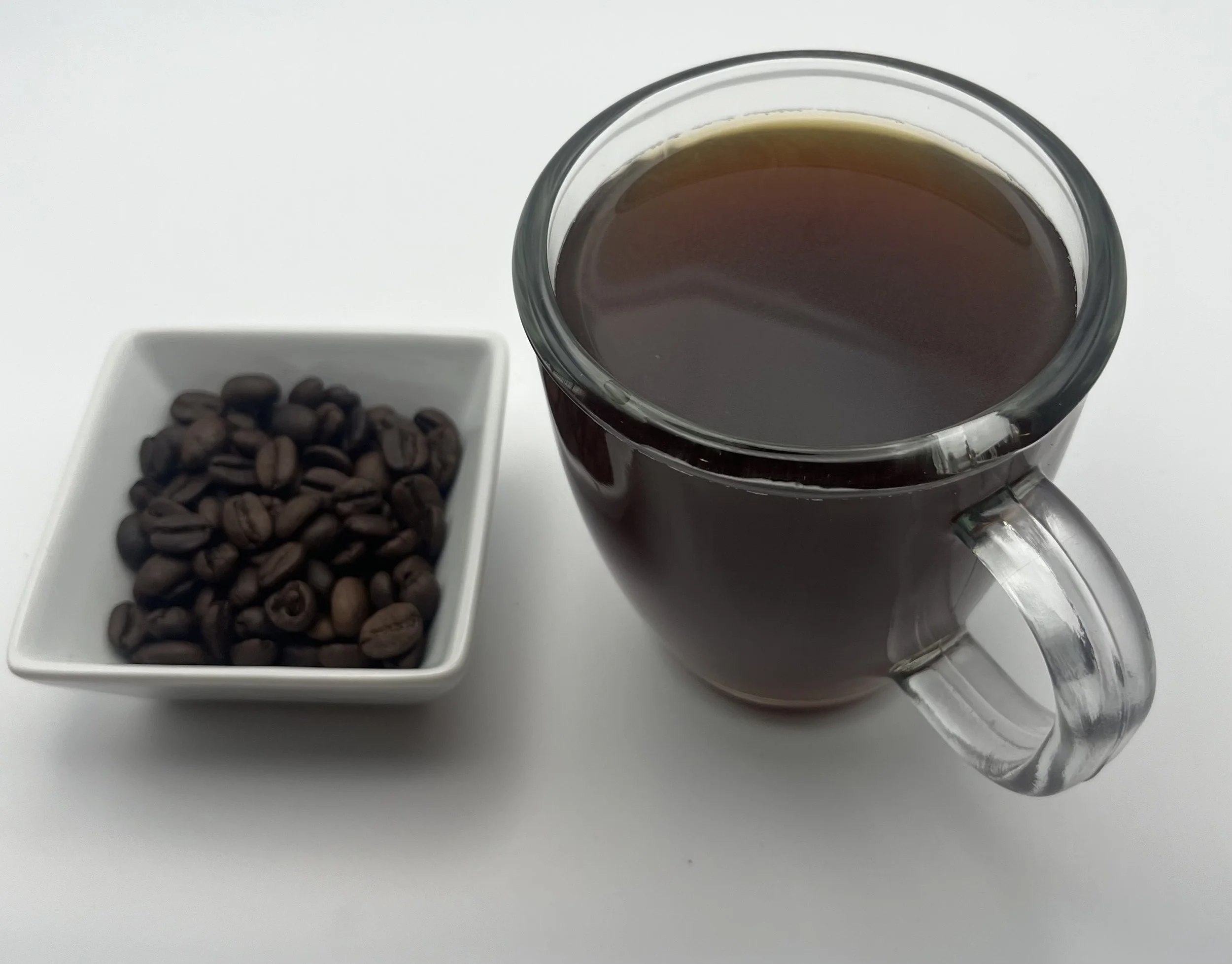 A white mug filled with black coffee, placed on a wooden surface with scattered coffee beans, with a blurred green outdoor background.
