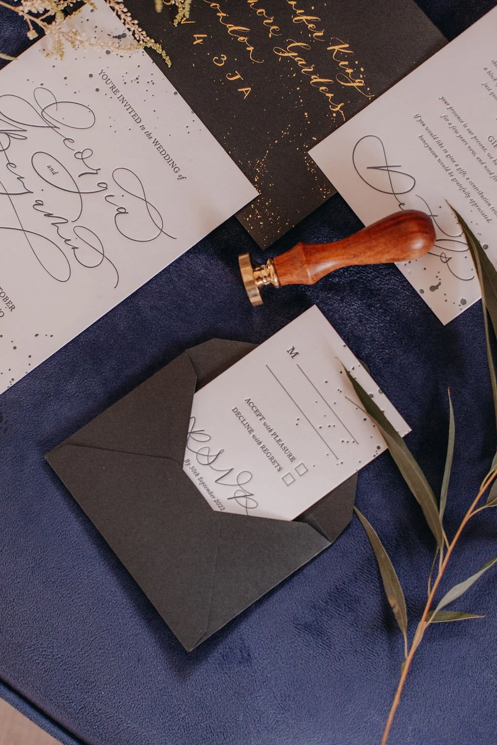 Wedding invitations and RSVP cards with black and white color scheme, gold and black decor, and a wooden wax seal stamp on a dark blue surface.