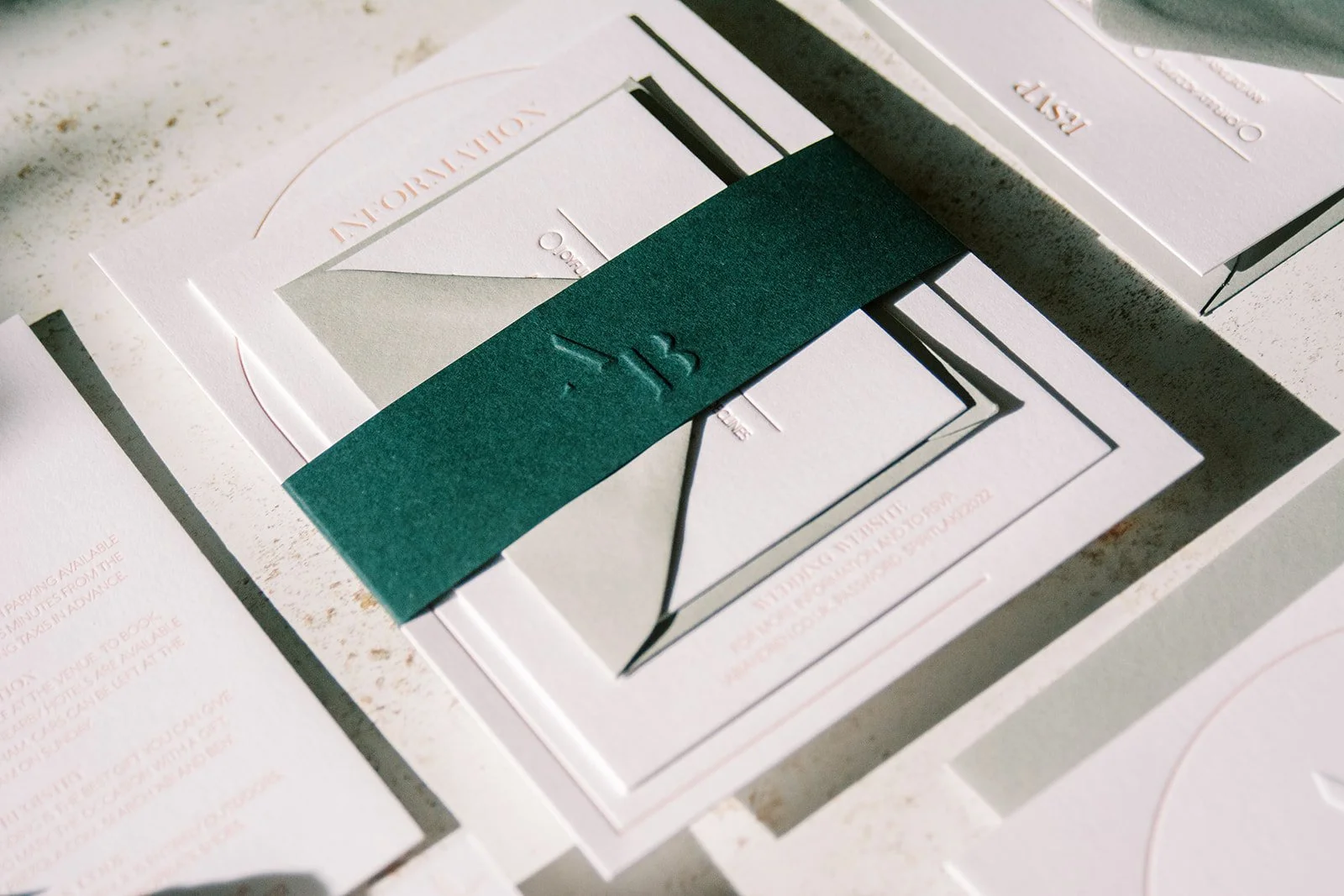 Close-up of elegant wedding invitations with a dark green ribbon embossed with initials A and B, on a light textured surface.