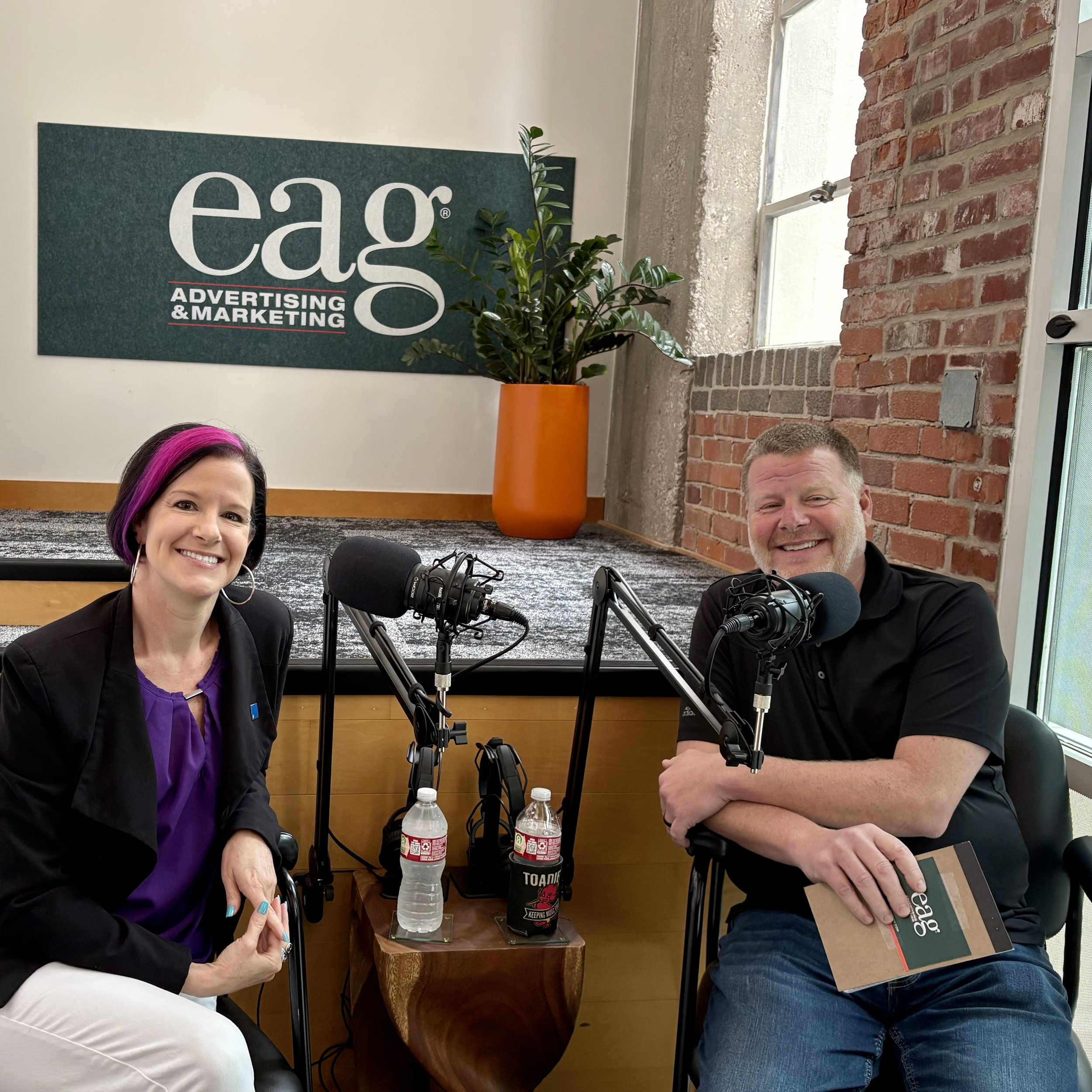 Photo of Julie Cortes and Jeff Randolph ready to record the podcast, sitting in front of large microphones with an EAG sign displayed behind them.