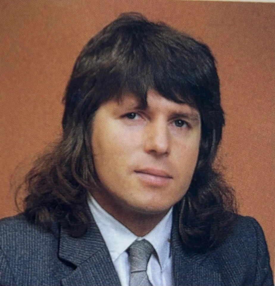 A man with long, dark hair wearing a gray suit and a white shirt.