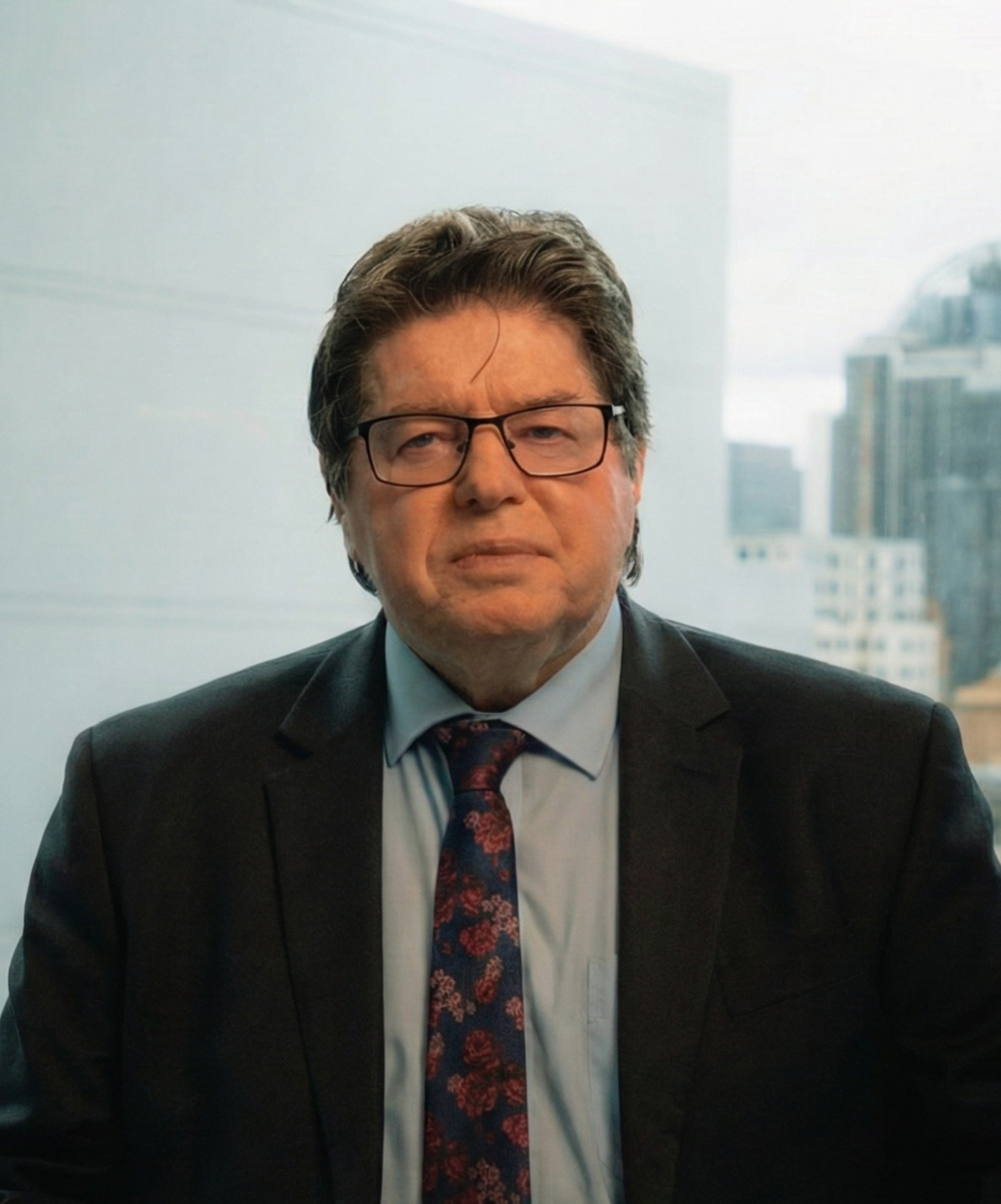 A man with dark hair, glasses, and a serious expression, wearing a suit, tie, and light blue shirt, sitting in an office with large windows showing a cityscape in the background.