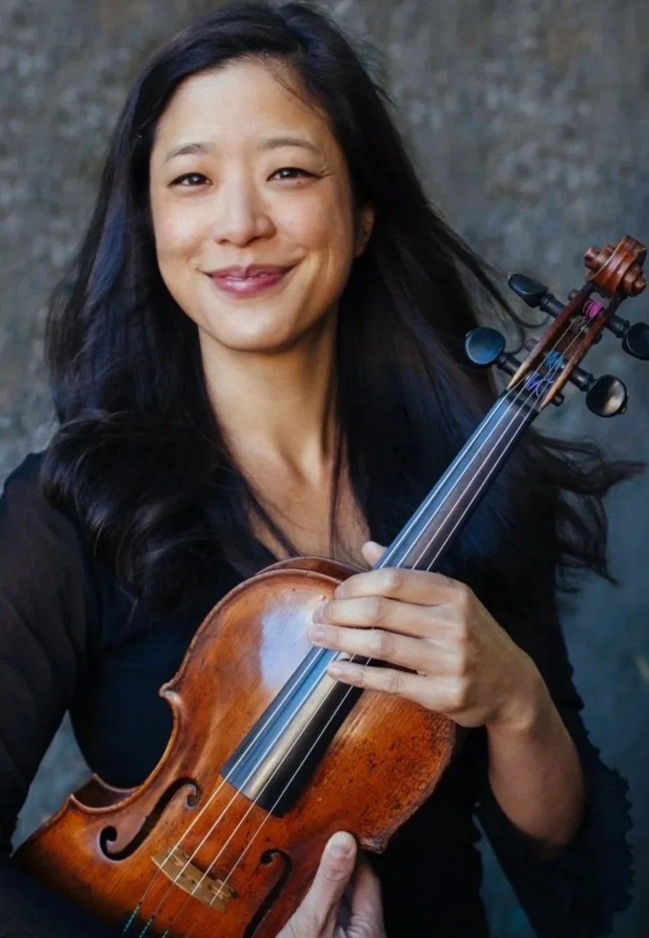 A smiling woman with long black hair holding a violin against a dark background.