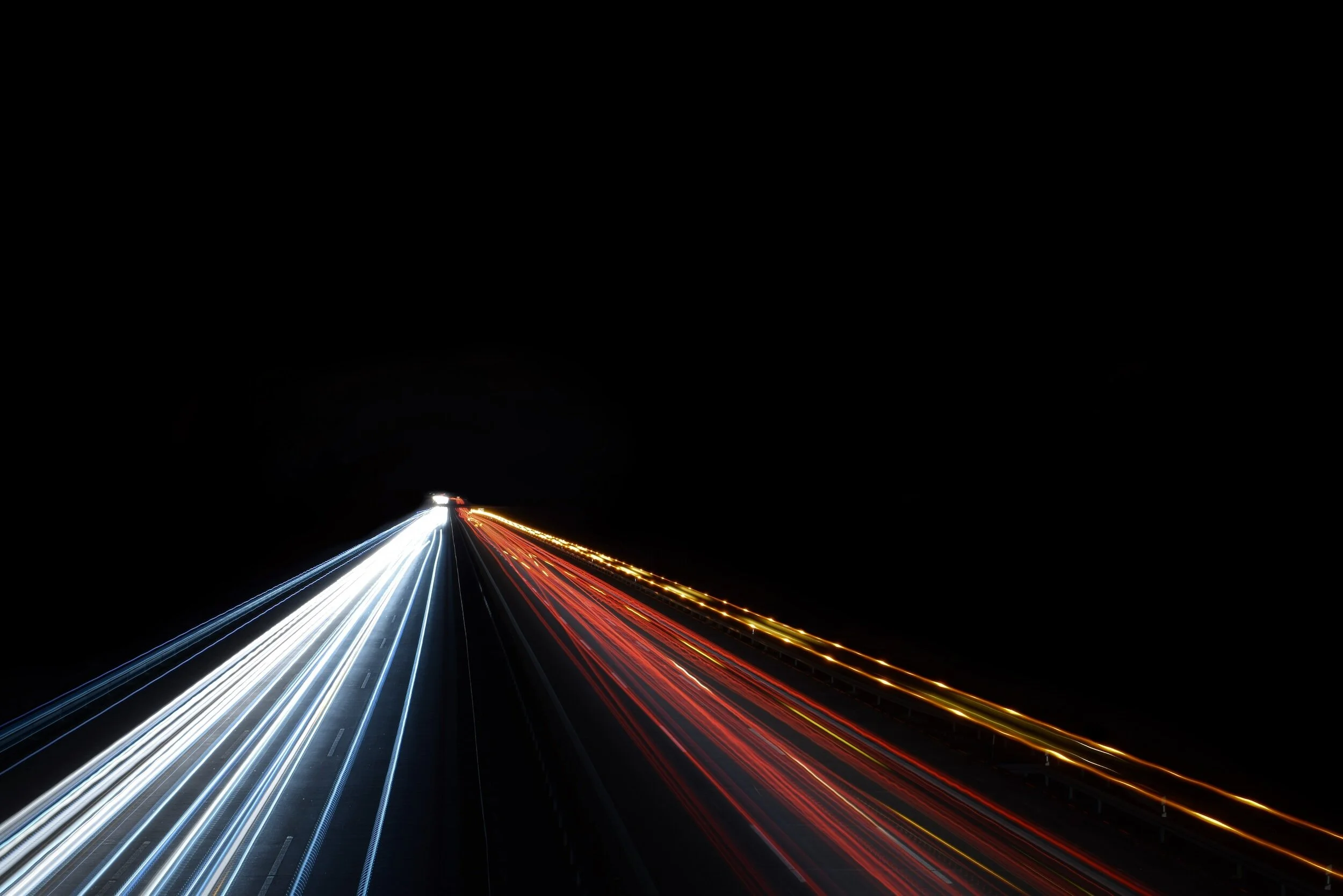 Long exposure photo of light trails on a highway at night, with white and blue lights moving in one direction and red and yellow lights moving in the opposite direction.