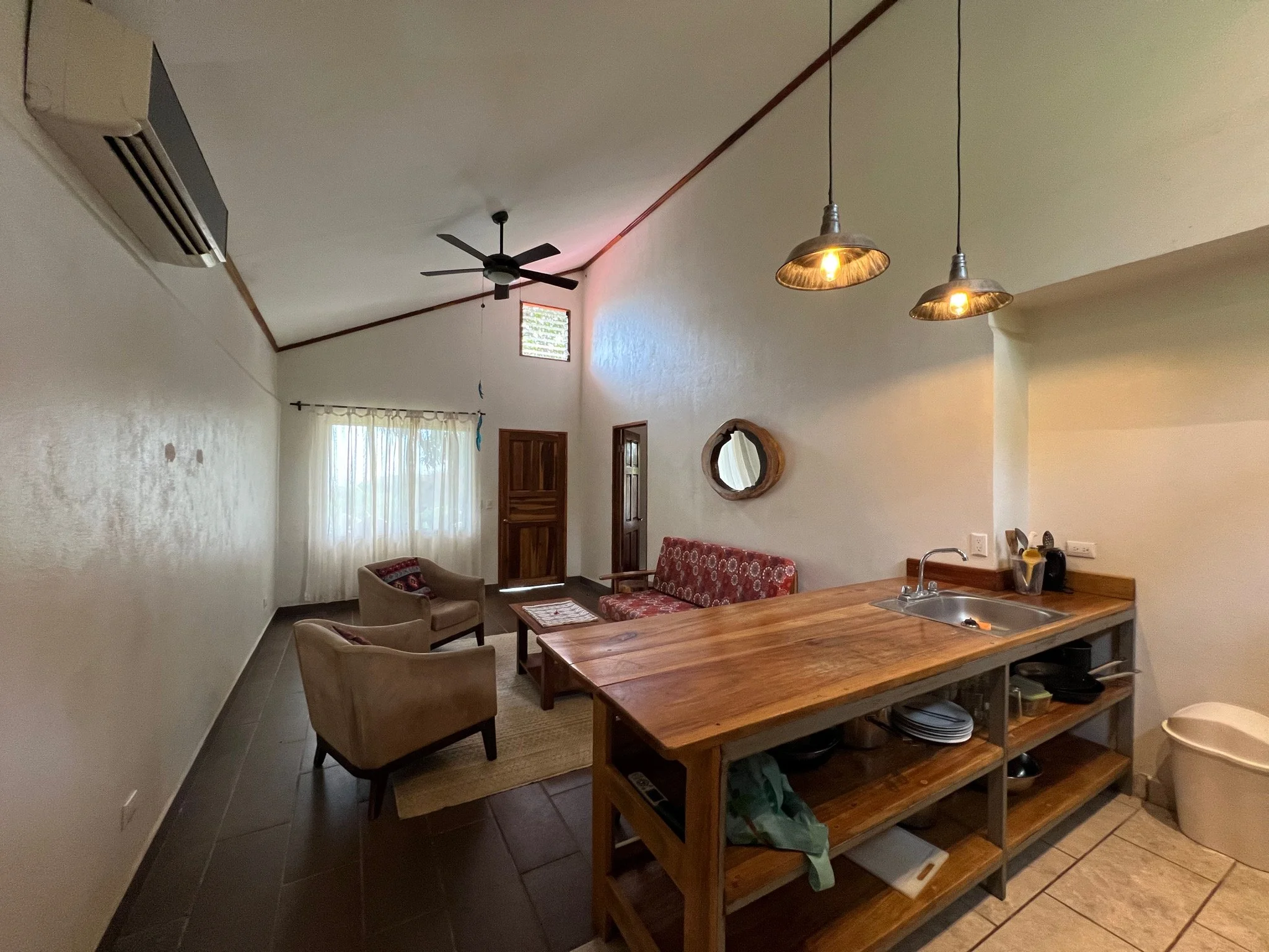 Living room with a wooden ceiling fan, a window with curtains, a small sitting area with two beige armchairs and a beige patterned sofa, and a kitchen counter with a sink, pendant lights, and various kitchen items.