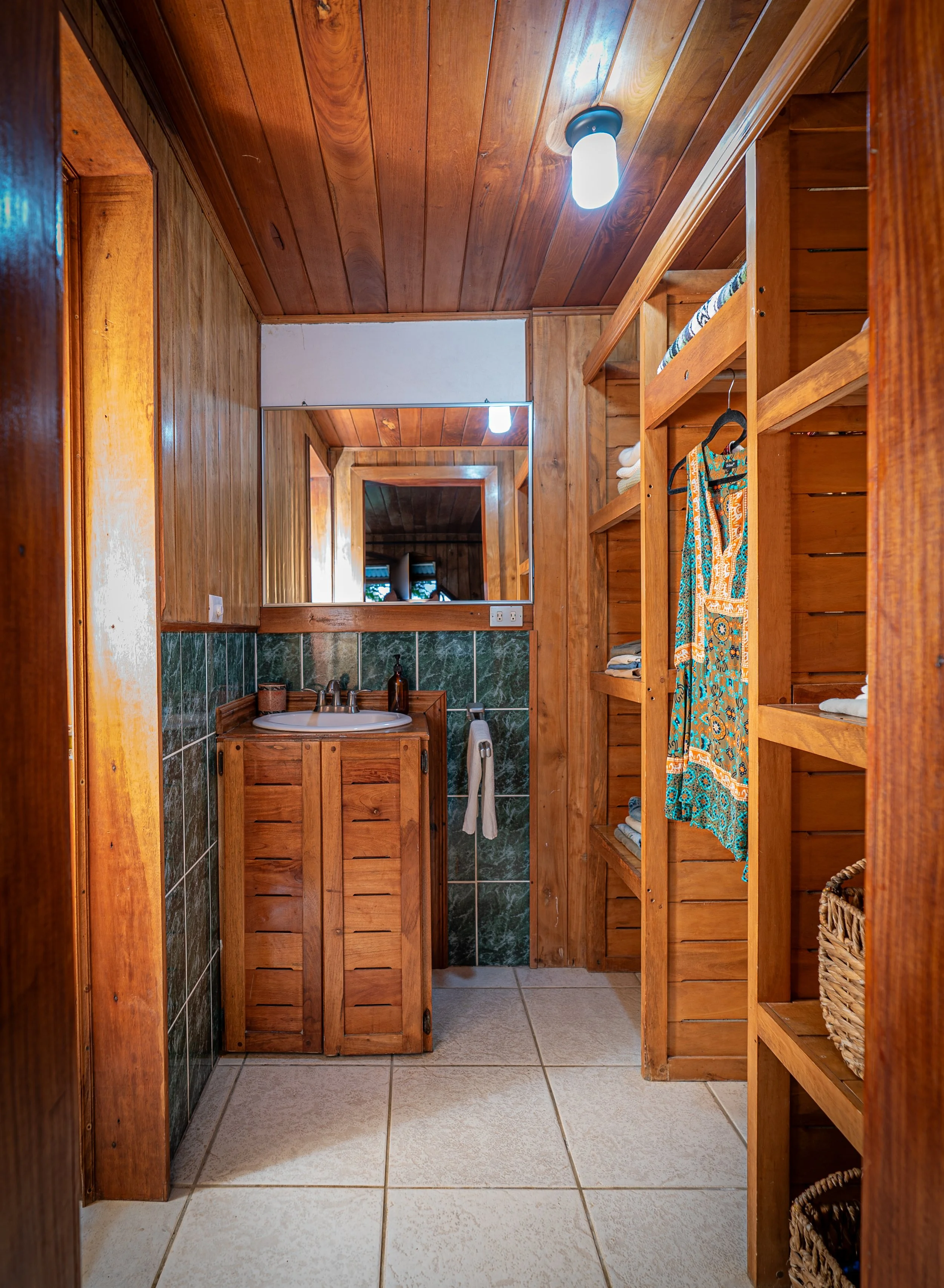 A rustic bathroom with wooden walls, ceiling, and shelves, featuring a mirror and small sink with a green tiled backsplash, and open shelving with towels and baskets.