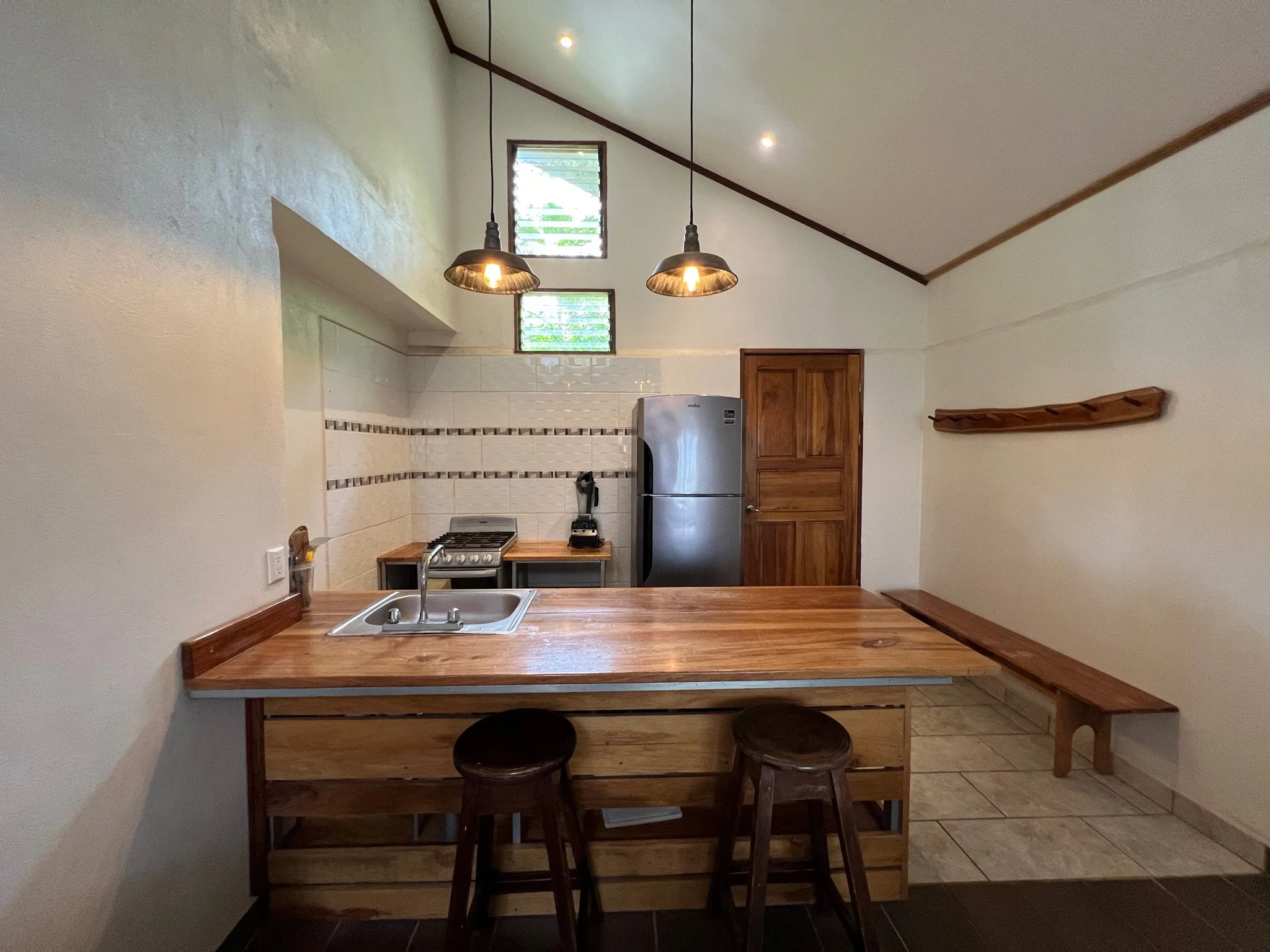 Kitchen with wooden island counter, two bar stools, a stainless steel refrigerator, a small stove, and a window with horizontal blinds, illuminated by pendant and ceiling lights.