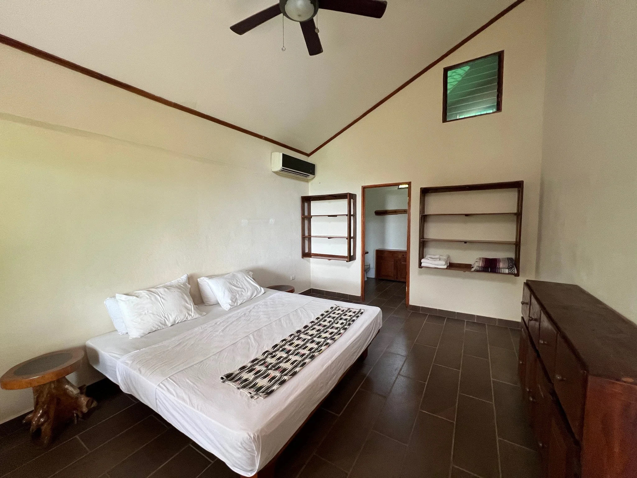 A bedroom with a bed, white bedding, and pillows, wooden side tables, wall-mounted shelves with towels, an air conditioning unit, a small window, and a ceiling fan, with an adjacent bathroom visible through an open door.