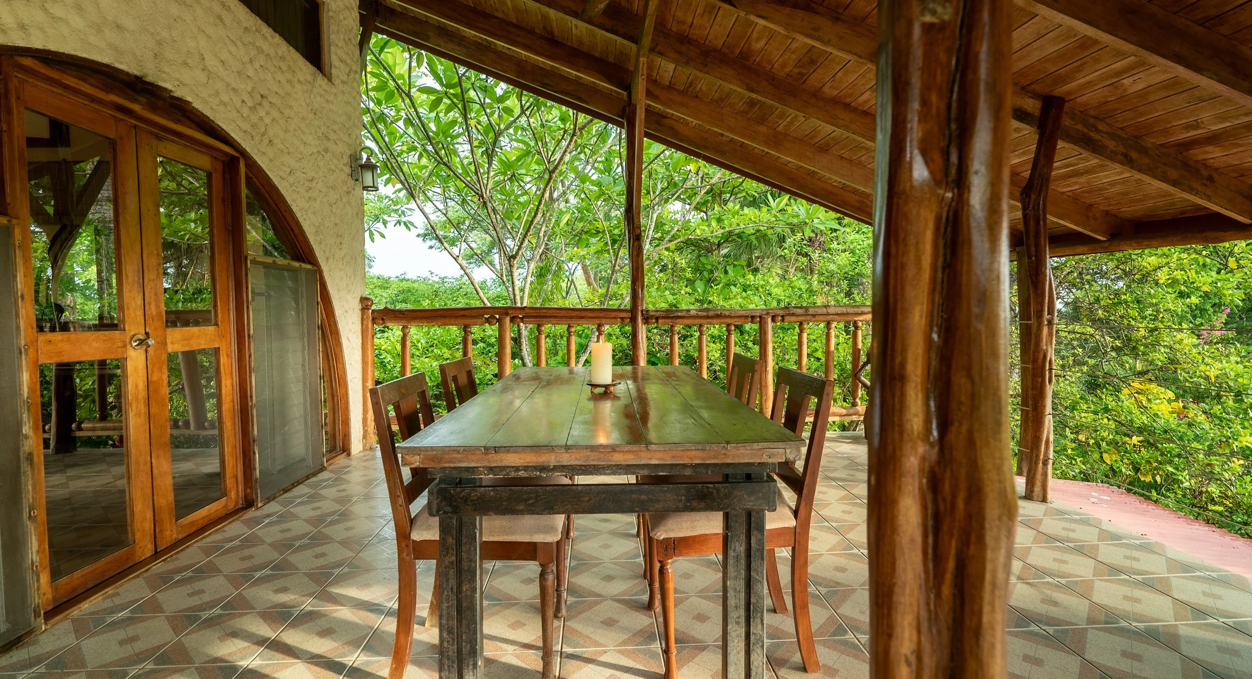 An outdoor wooden dining area with a large table, six chairs, a lit candle on the table, surrounded by lush green trees and a wooden railing.