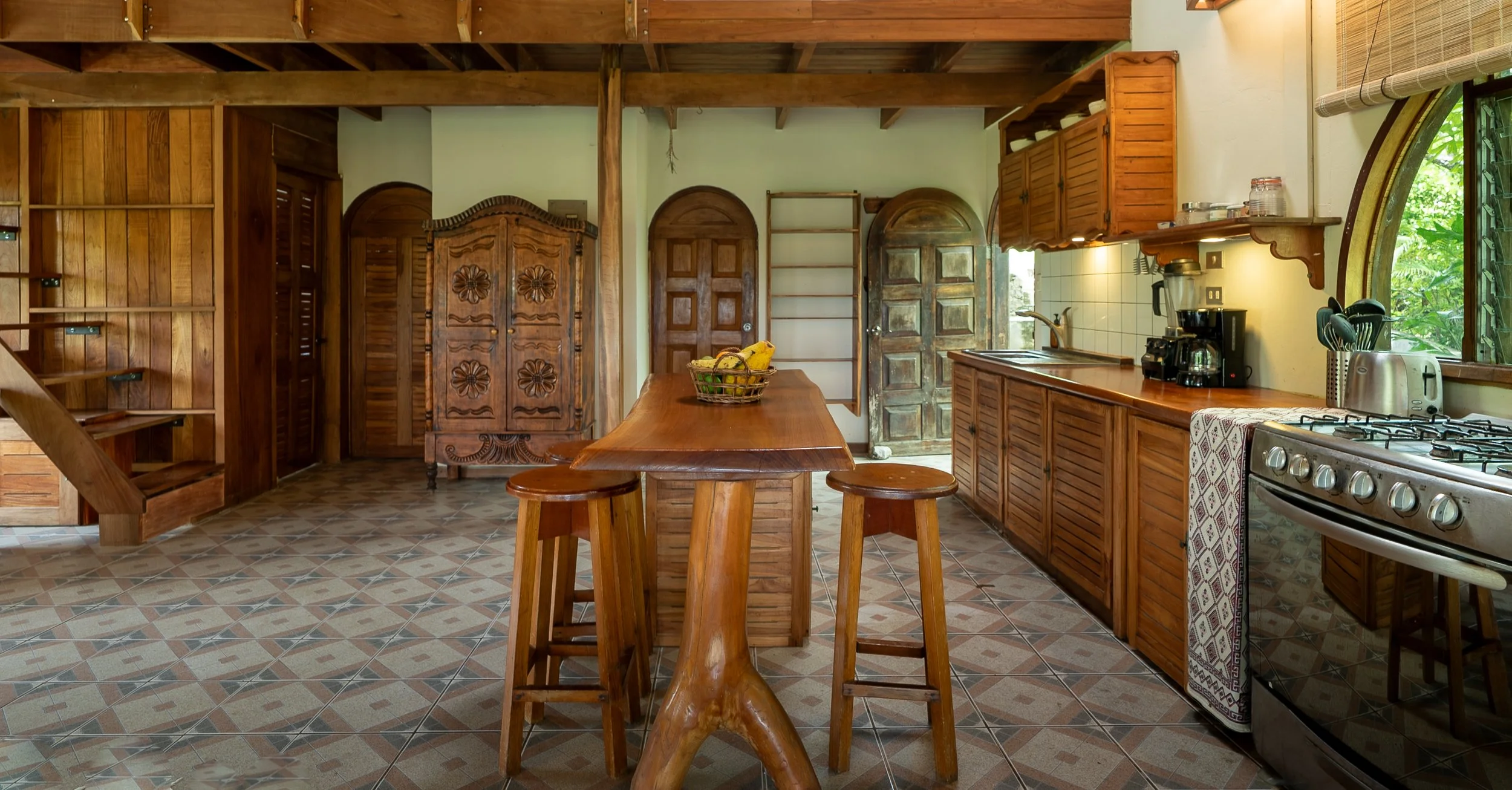Kitchen with wooden cabinets, a central wooden table with stools, a fruit basket, a stove, coffee maker, toaster, and a window with a view of greenery.