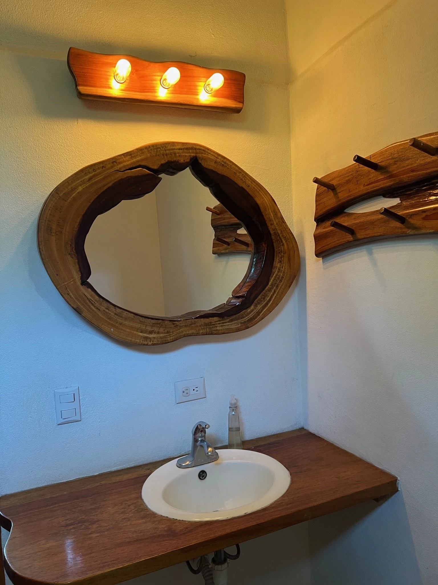A rustic bathroom vanity with a wooden countertop, a small sink, and a handheld soap dispenser. Above the mirror, a wooden light fixture with three exposed bulbs provides illumination. A decorative mirror with an irregular wooden frame hangs on the wall, along with two wooden wall-mounted racks to the right.