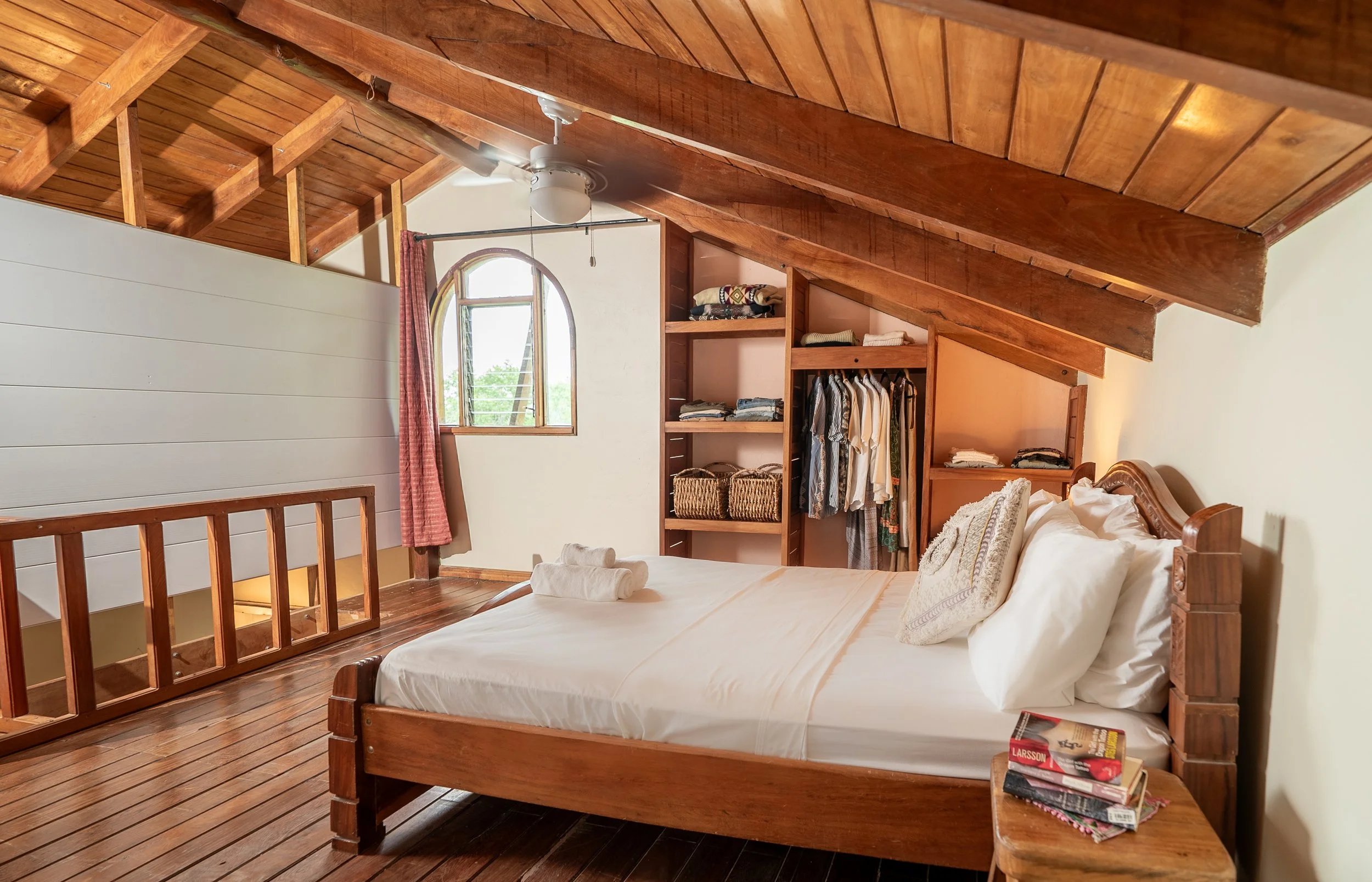 Cozy bedroom with wooden ceiling and floors, a bed with white linens and pillows, an open closet with clothes and baskets, a small bedside table with books, a window with an arched top and pink curtains, and a ceiling fan.